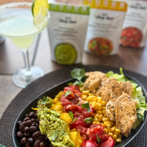 Tacos on a black plate with black beans, corn, diced tomatoes, and guacamole, accompanied by a green cocktail with a lime wedge, on a wooden table with packaging of seasoning mixes in the background.