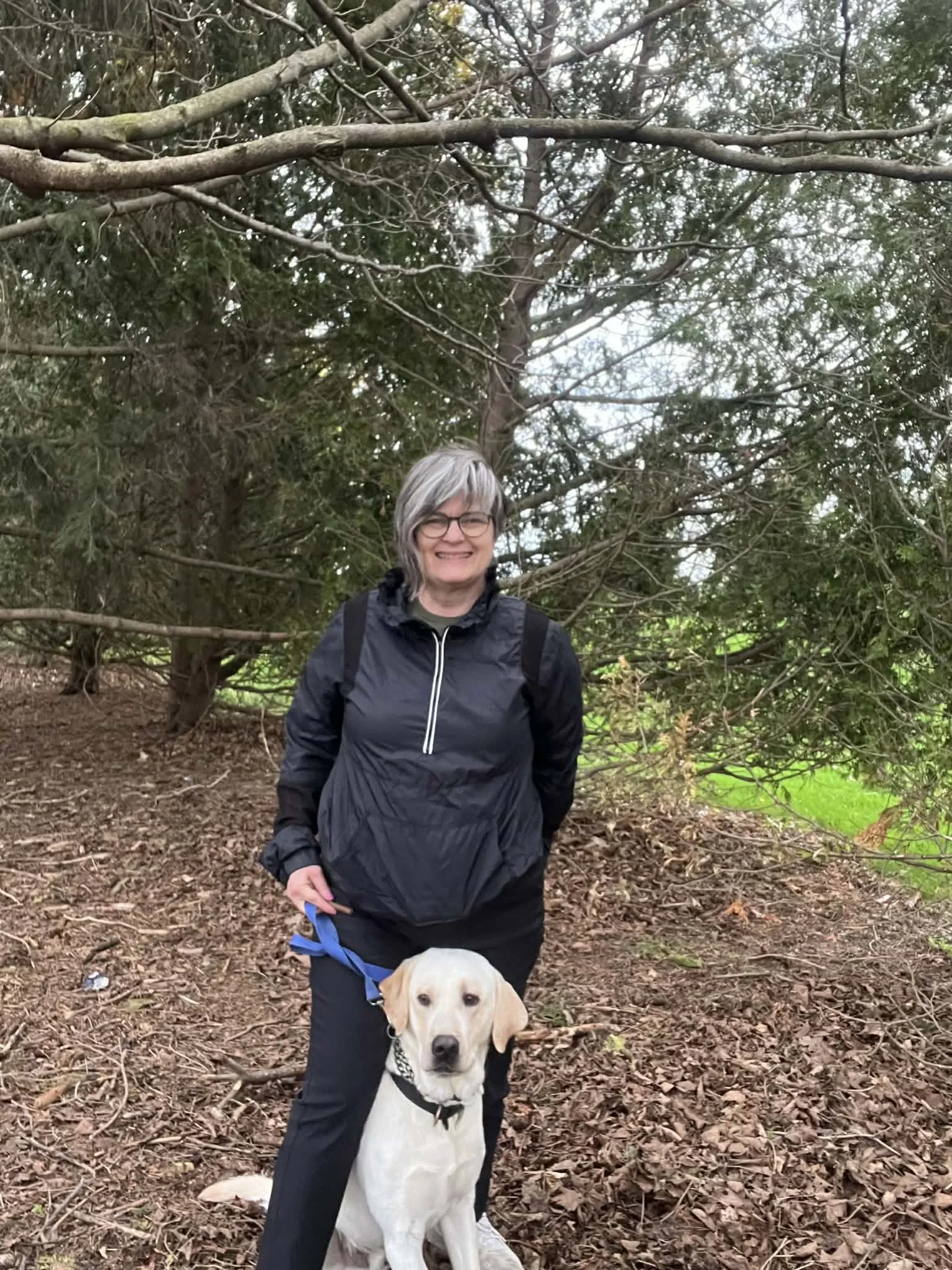 A smiling woman with glasses and gray hair stands in a forest with a large, white dog. The woman is wearing a black jacket and has a backpack, while the dog, sitting obediently, has a black collar and leash. The forest background includes trees, branches, and a ground covered with leaves.