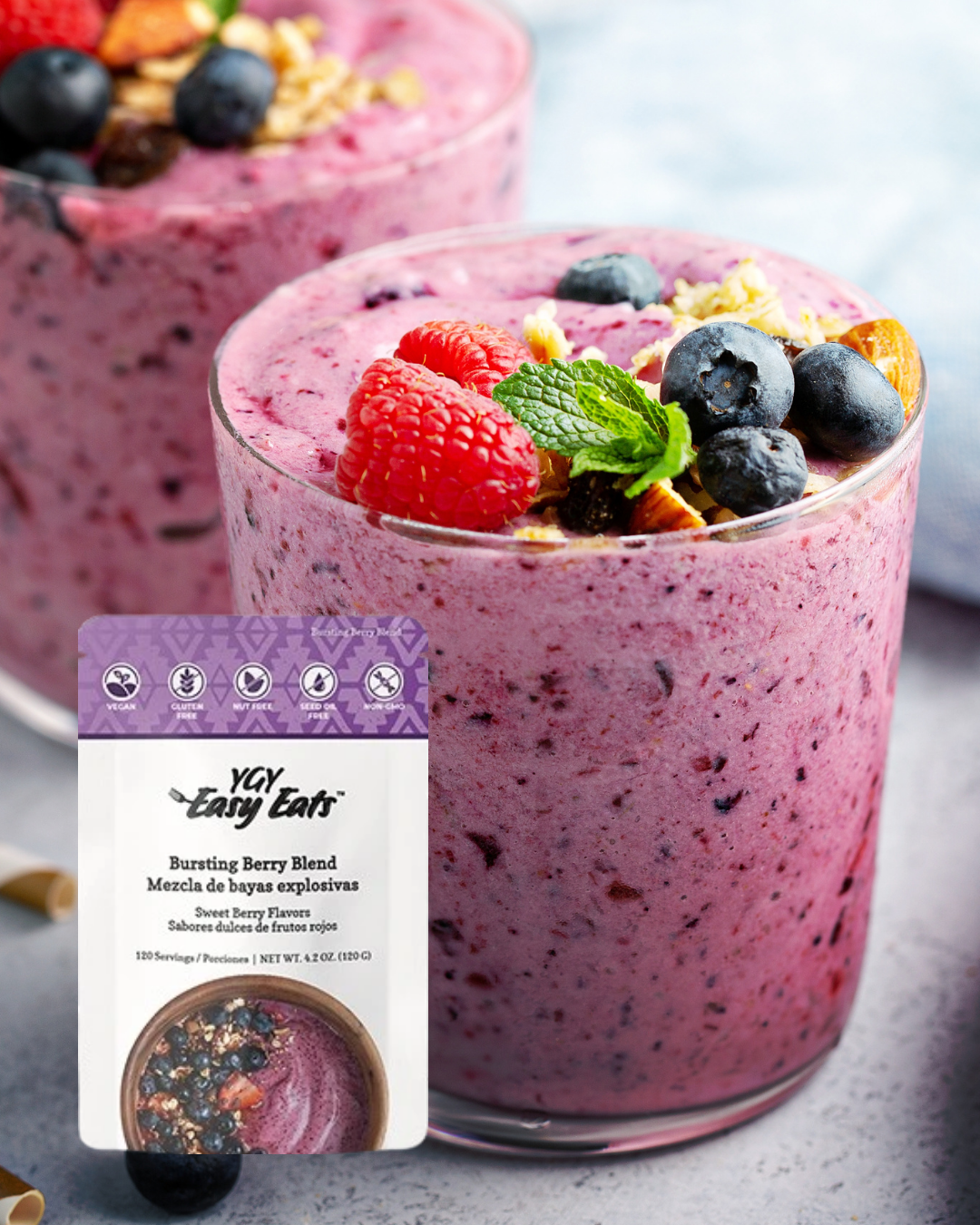 Two glasses of berry smoothie topped with fresh raspberries, blueberries, a mint leaf, and granola, with a package of YAY Easy Eat bursting berry blend mix in the foreground.