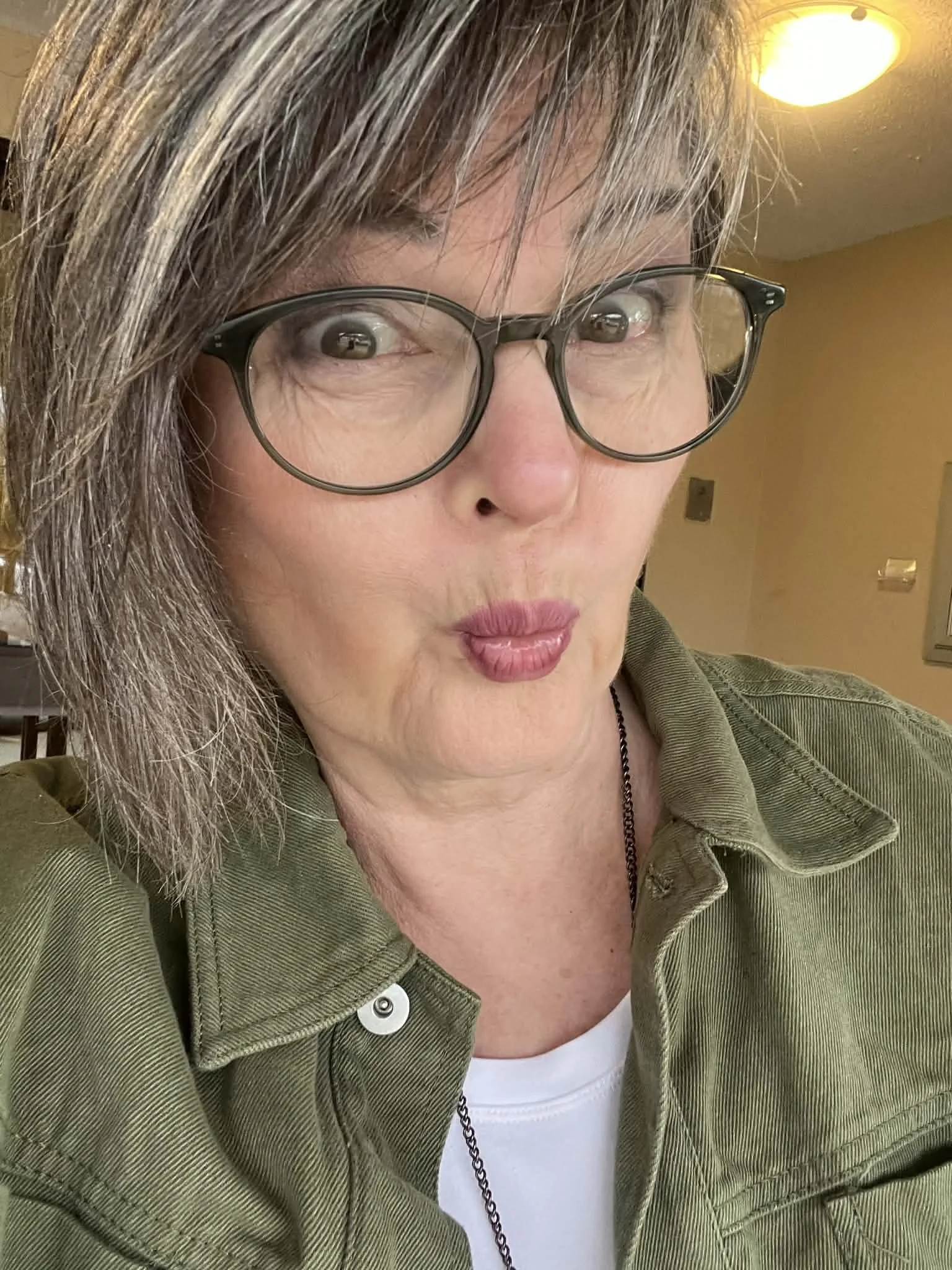 Close-up of a woman with gray hair wearing large glasses, a green jacket, and a white shirt, making a playful face indoors with a yellow ceiling light above.