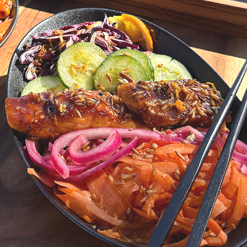 A black bowl containing grilled chicken, pickled carrots, pickled onions, sliced cucumbers, red cabbage slaw, and a lemon wedge, with chopsticks resting on the bowl's edge.