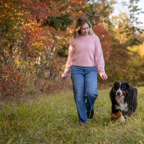 A woman walking with a dog in a grassy field during fall, with colorful autumn trees in the background.