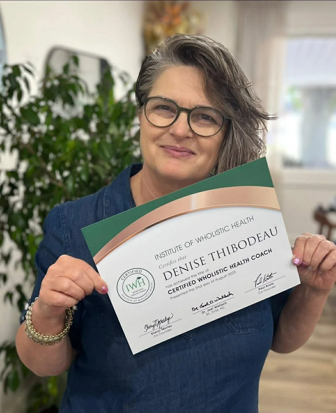 A smiling woman with short brown hair, glasses, and a denim shirt, holding a certificate from the Institute of Wholistic Health.