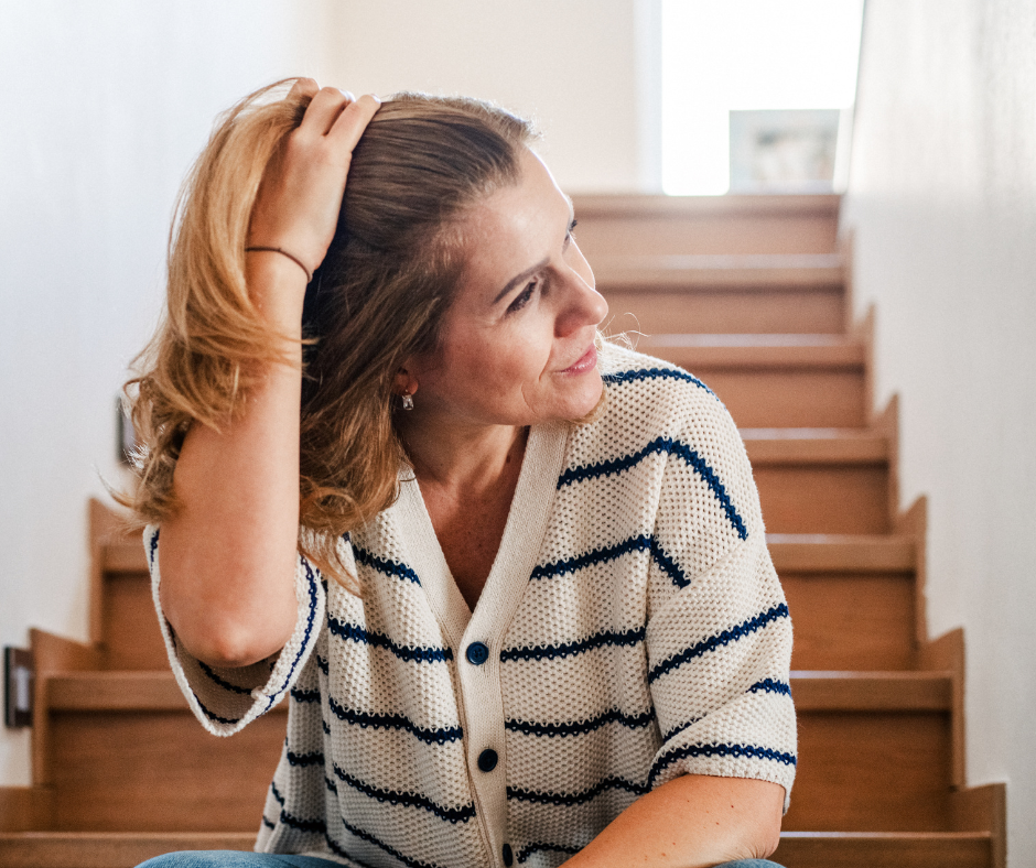 A woman with shoulder-length, wavy hair sitting on wooden stairs, wearing a white and navy striped cardigan, touching her hair, looking to the side with a slight smile.
