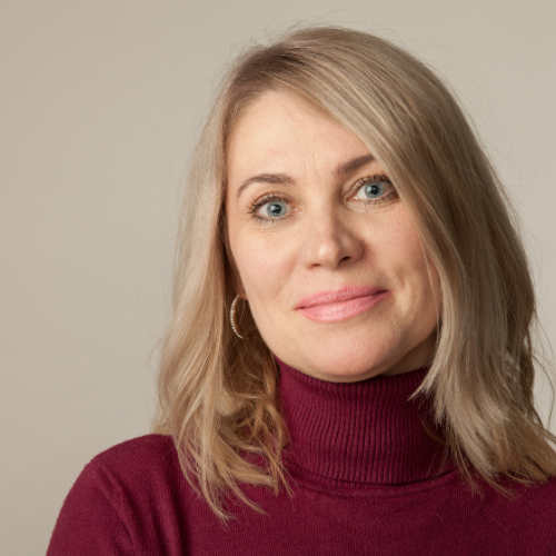 Portrait of a woman with blonde hair and blue eyes wearing a burgundy turtleneck sweater and hoop earrings, smiling softly against a neutral background.
