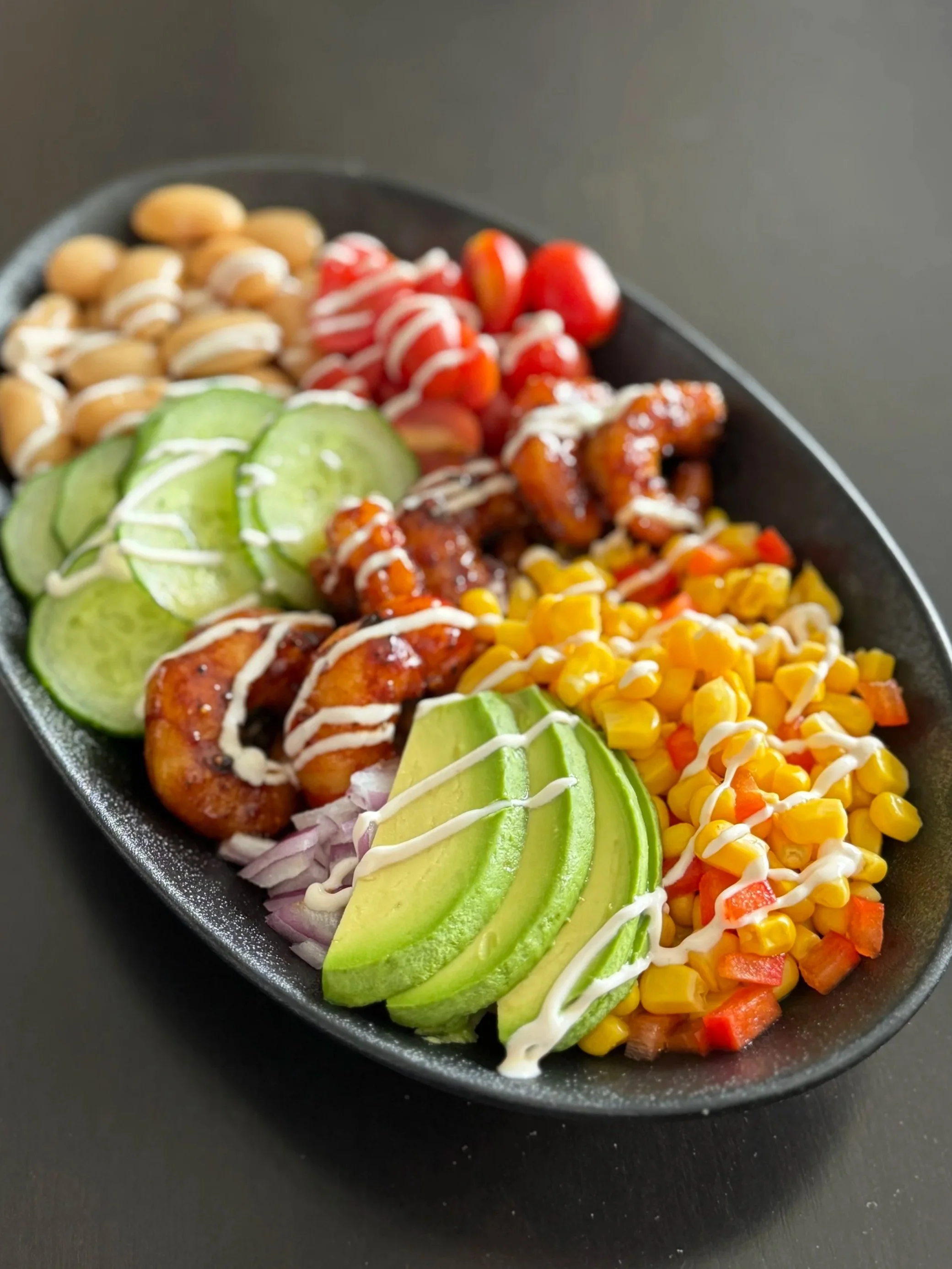 A black oval dish containing sliced avocado, cherry tomatoes, grilled chicken pieces, corn, diced red bell peppers, and sliced cucumbers, all drizzled with white dressing.