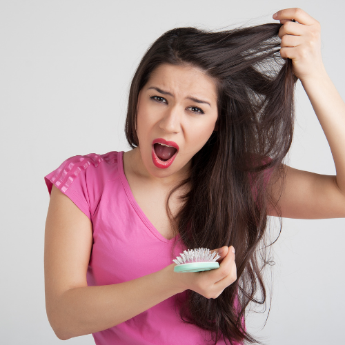 Young woman with long dark hair, wearing a pink shirt, holding a hairbrush, pulling her hair and making a frustrated expression.