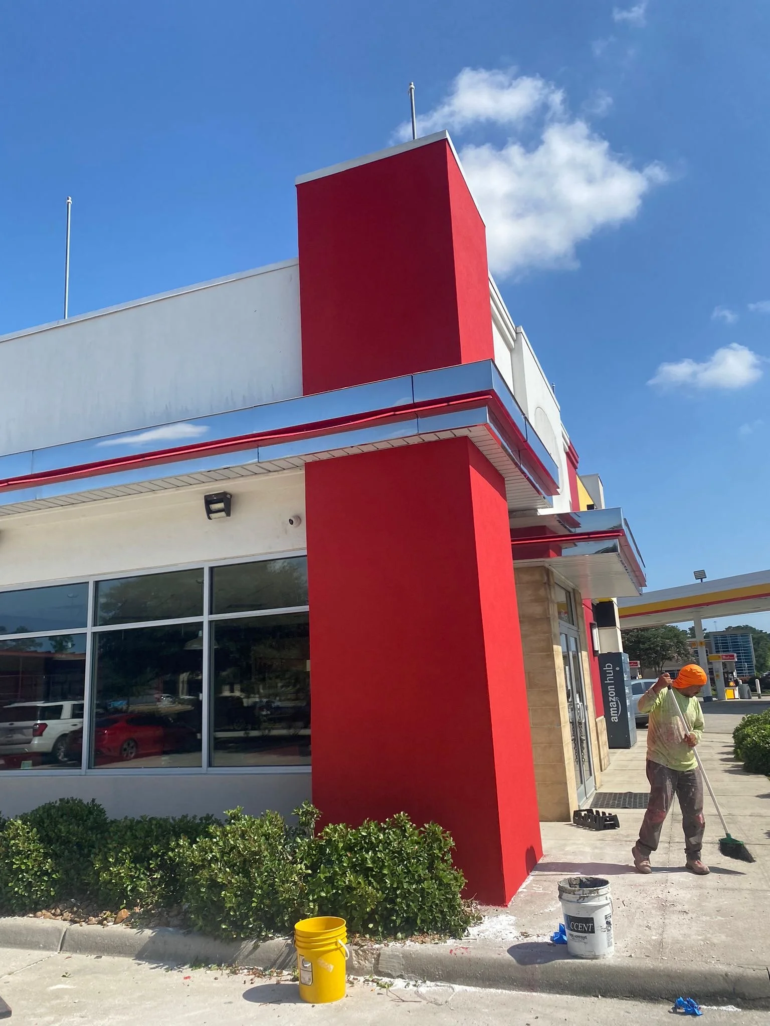 A worker with an orange hat sweeping the sidewalk outside a modern building with red and white exterior walls, large glass windows, and a gas station in the background.