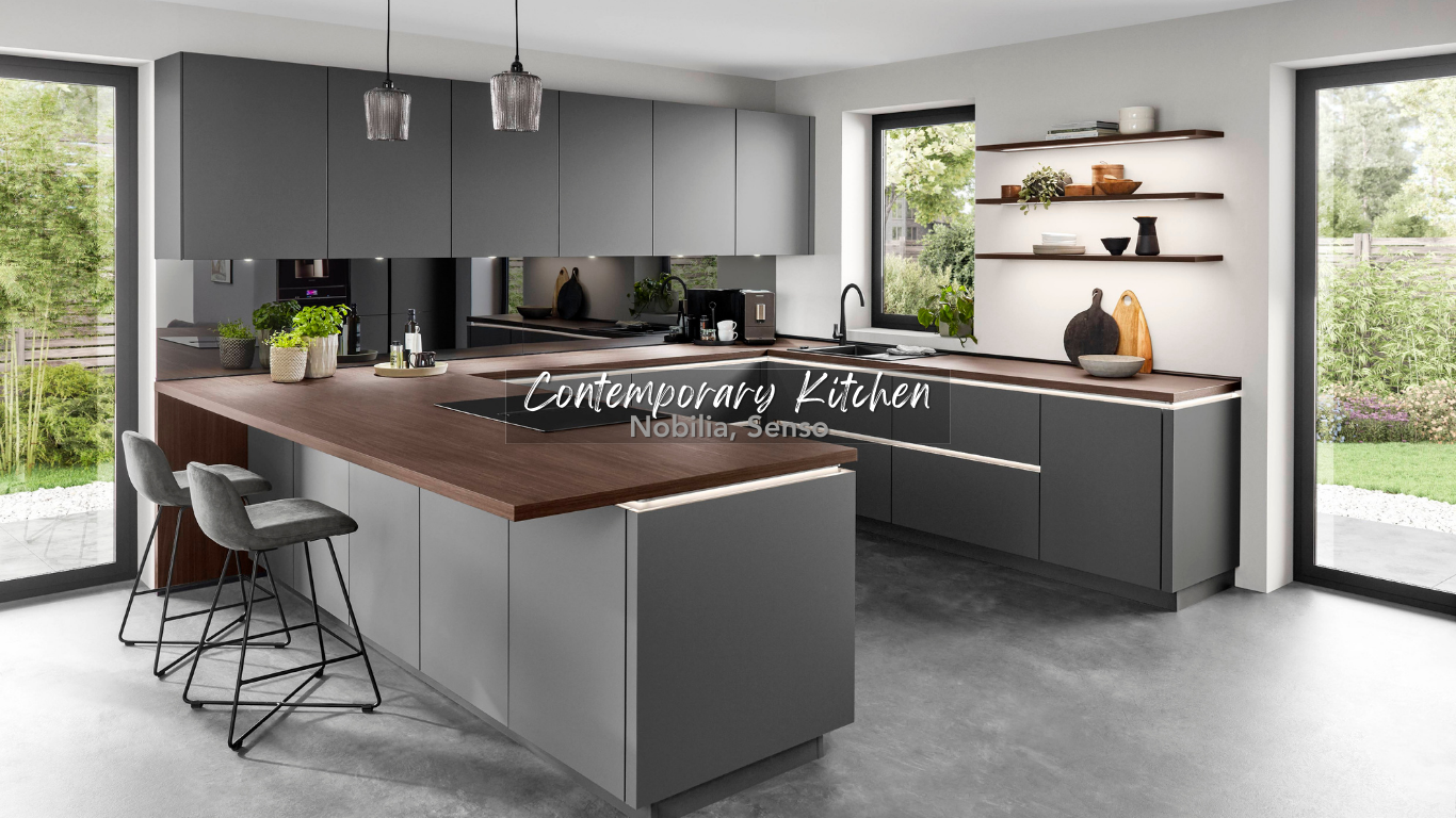 Modern kitchen with gray cabinets, wooden countertop island, two bar stools, large windows showing a backyard with greenery, and open shelves with decor.