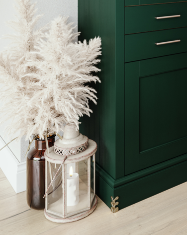 Decorative white pampas grass in a brown glass vase, with a candle inside a glass lantern, next to a green cabinet with drawers, in a well-lit room.