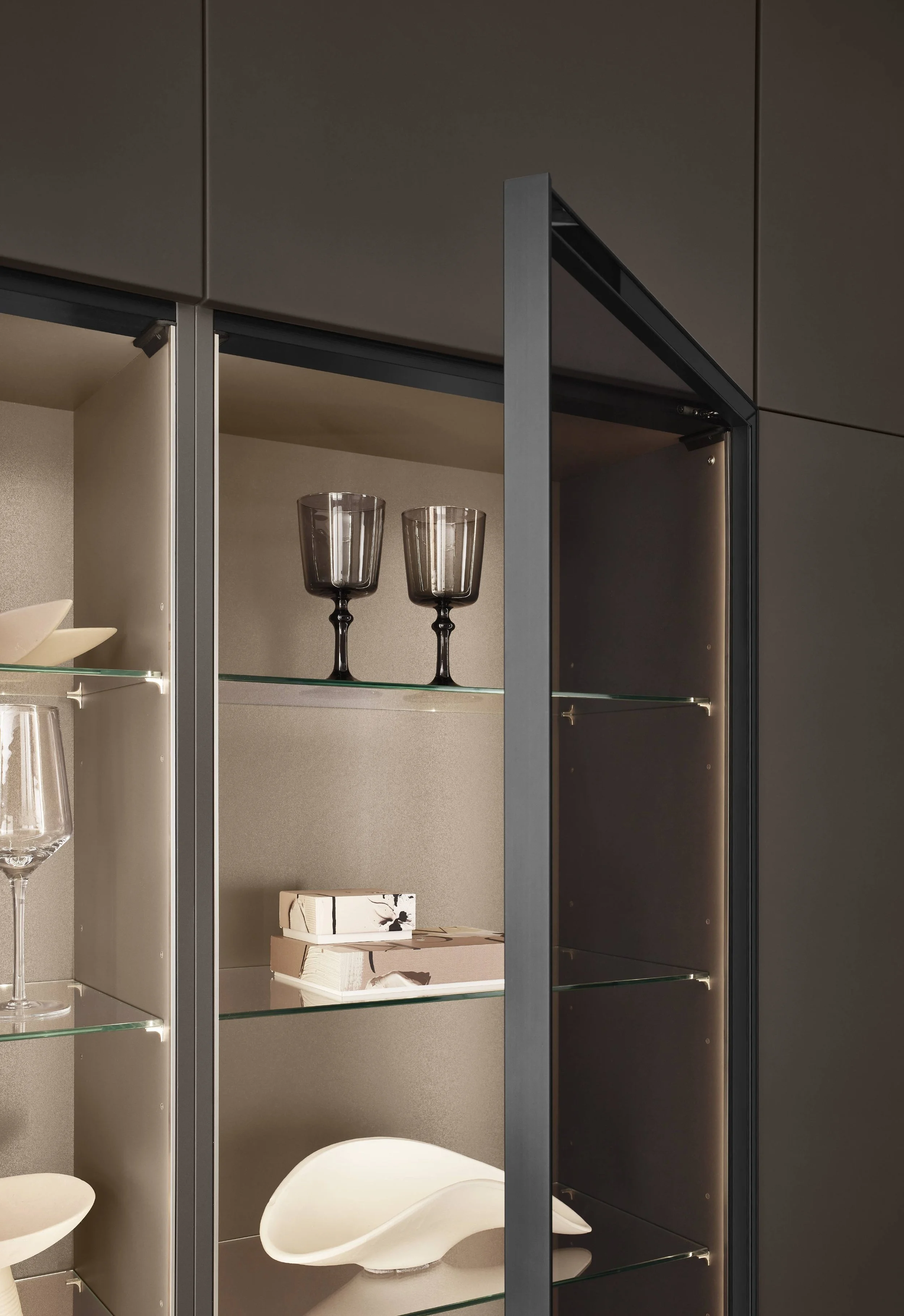 Close-up of a modern glass display cabinet with black frame, containing wine glasses, decorative boxes, and a white ceramic object, set against a brown wall.