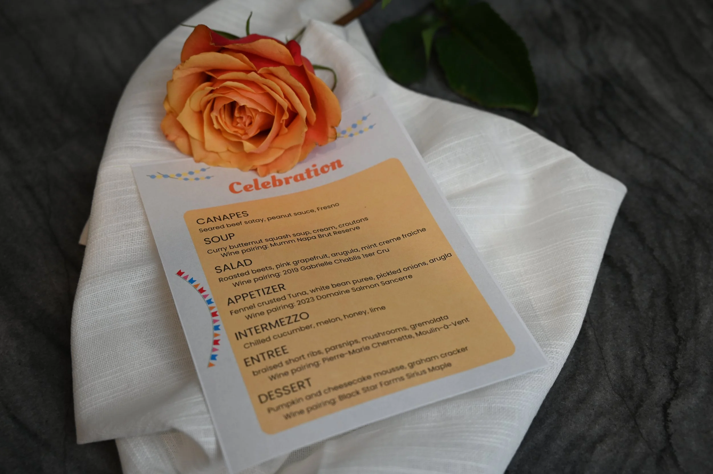A wedding menu on a white cloth with a peach-colored rose on top. The menu features various dishes and wine pairings, titled 'Celebration'.