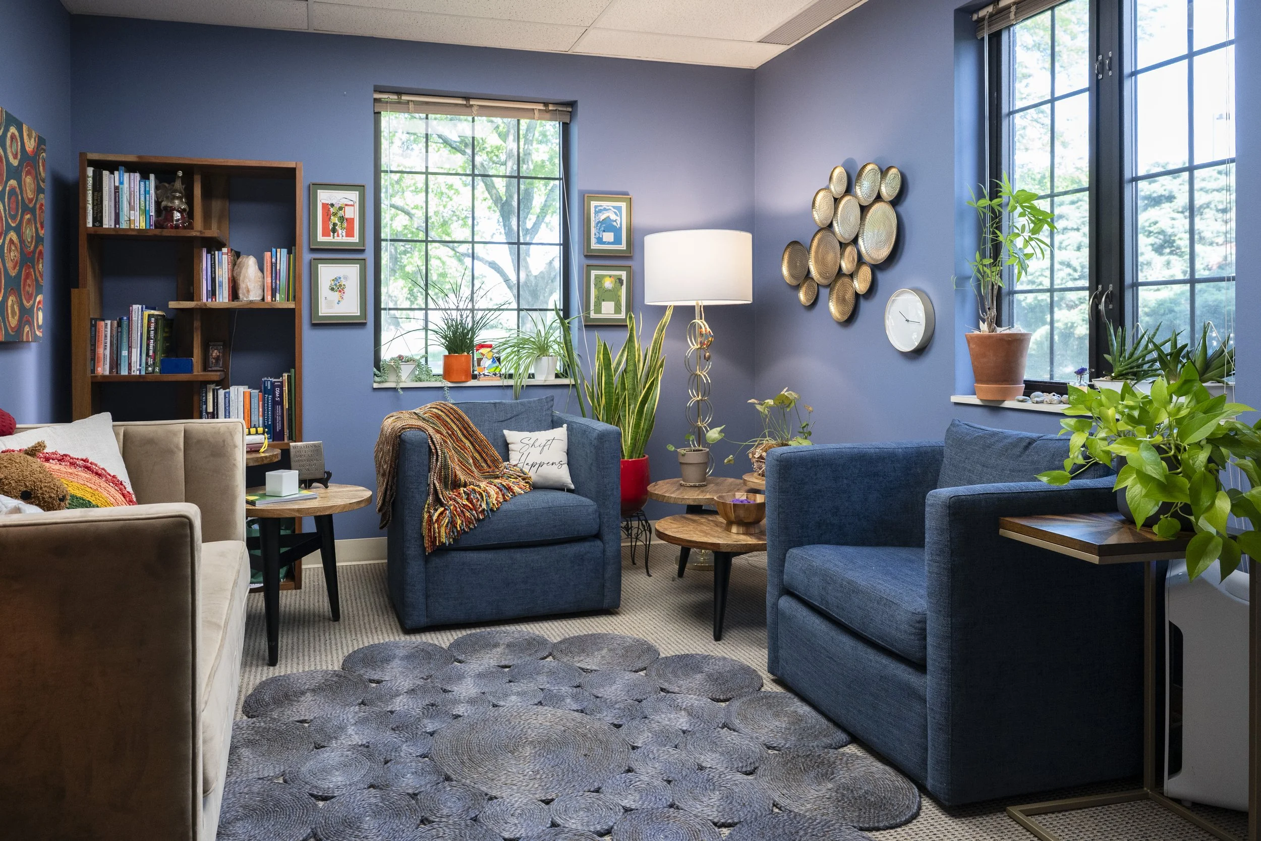 A cozy living room with blue painted walls, large windows showing green trees outside, and various plants on the windowsill and furniture. The room features a mix of furniture including a beige sofa, blue armchairs, and small wooden tables, accented with colorful artwork, decorative plates, a tall floor lamp, and a rug with a circular pattern.