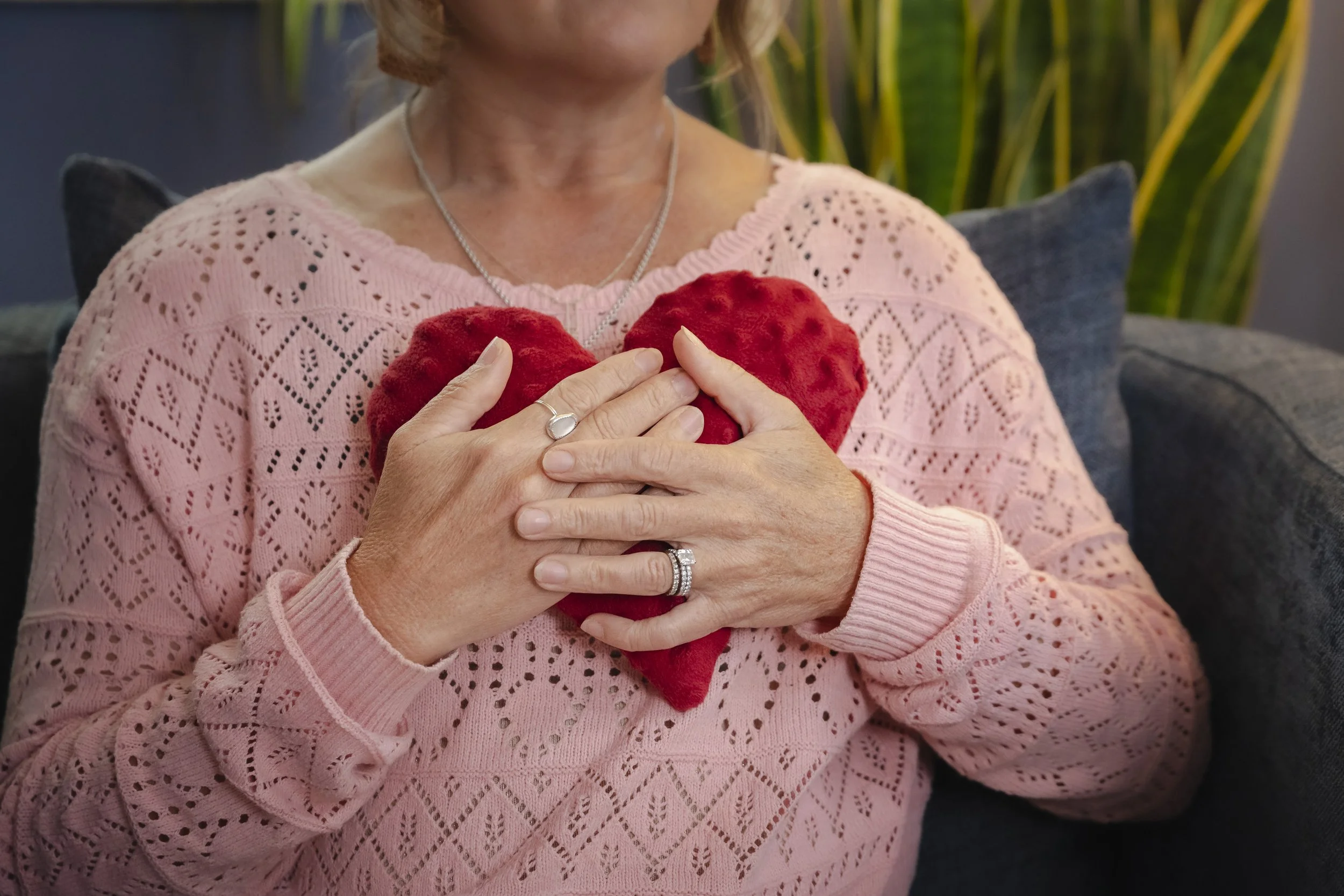 A woman in a therapy session wearing a pink sweater holding a plush red heart close to her chest with both hands. She has a silver ring on her left hand and a silver necklace. The background includes green plants and a gray cushion.