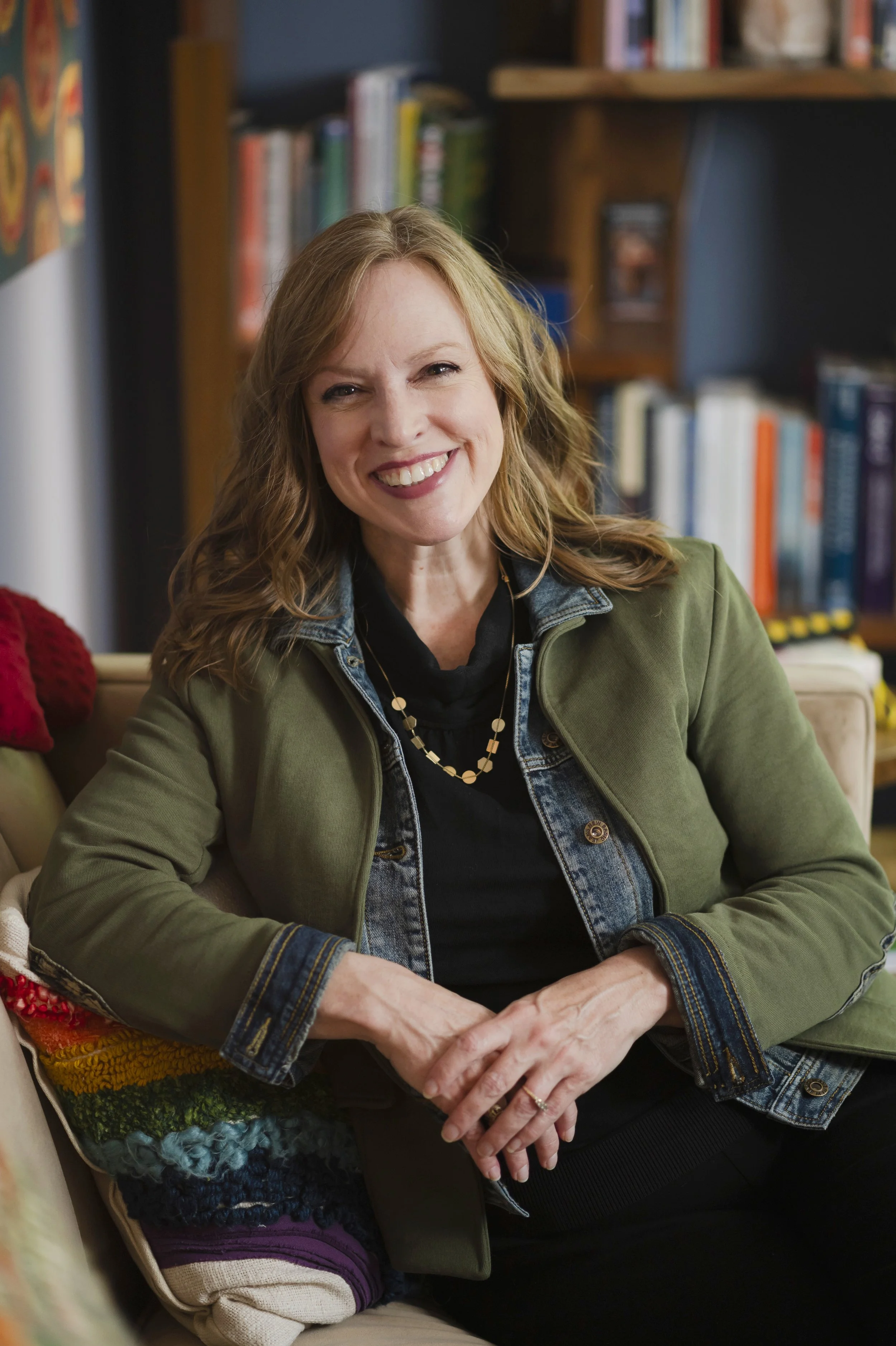 A smiling somatic experiencing practitioner with wavy red hair, wearing a green jacket, black shirt, and layered necklaces, sitting in a therapy office with bookshelves in the background.