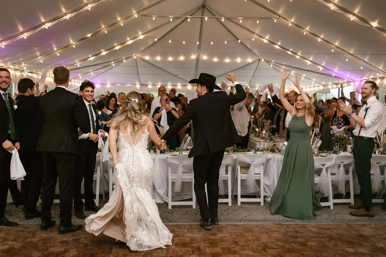  A bride and groom walking into their reception in a large white tent and lights above them, with wedding guests cheering them on. 