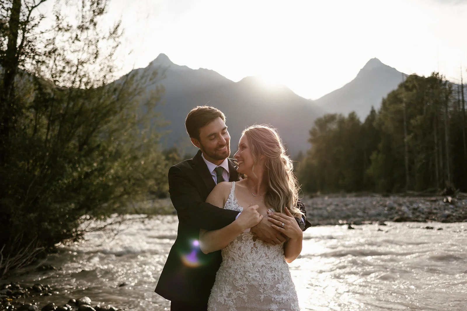  A bride and groom hugging each other with mountains and a river behind them at Cheekye Ranch. 