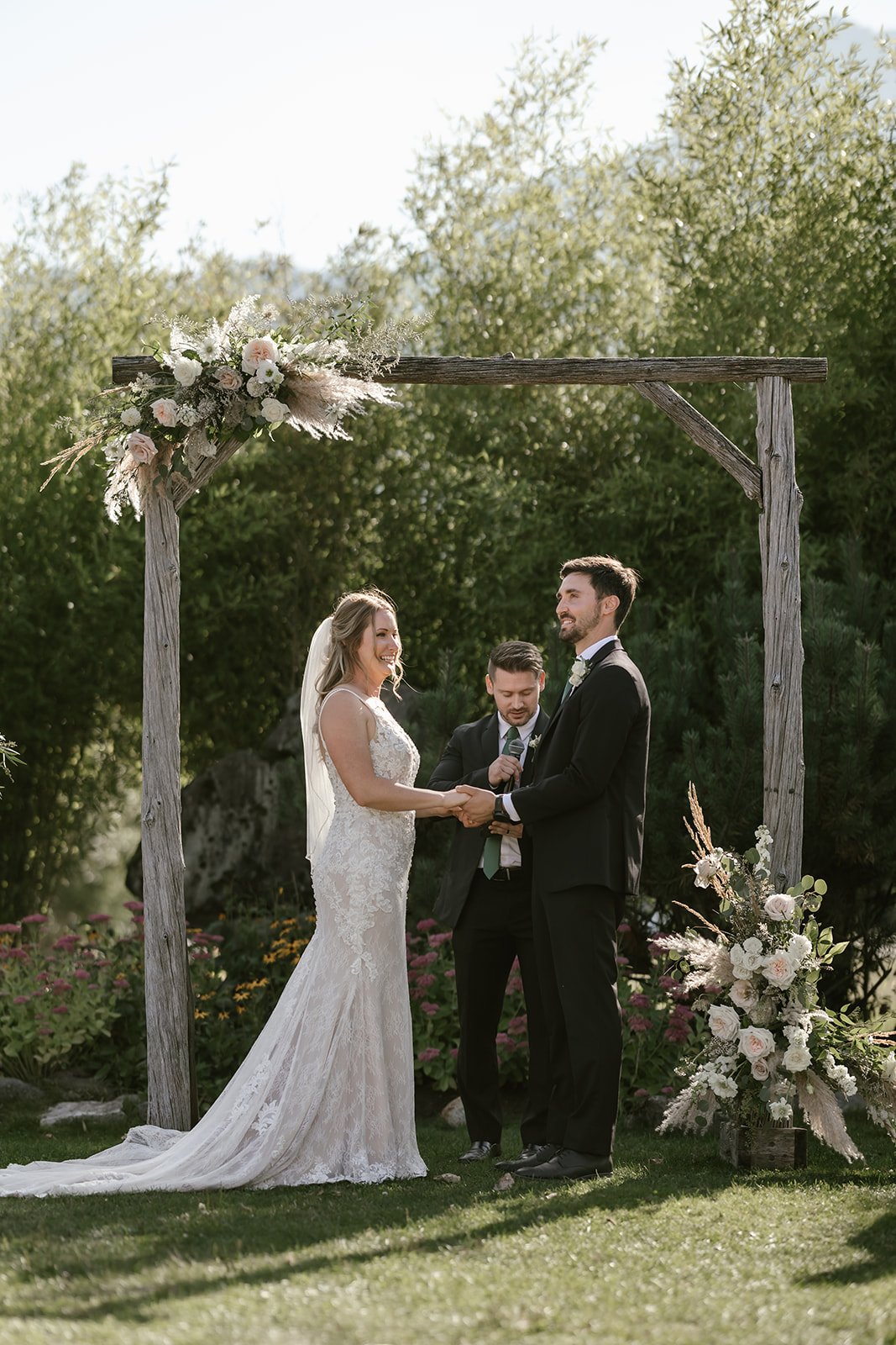  A bride and groom standing before a structure for their wedding ceremony with greenery and trees behind them.  