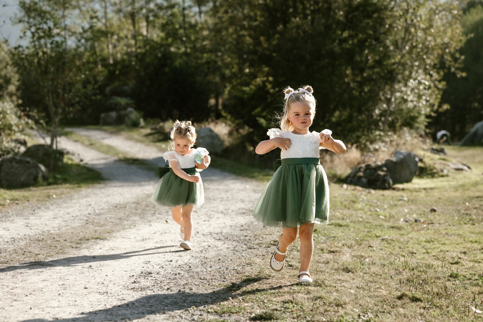  Two small girls running along a path in green skirts and white tops with trees behind them.  