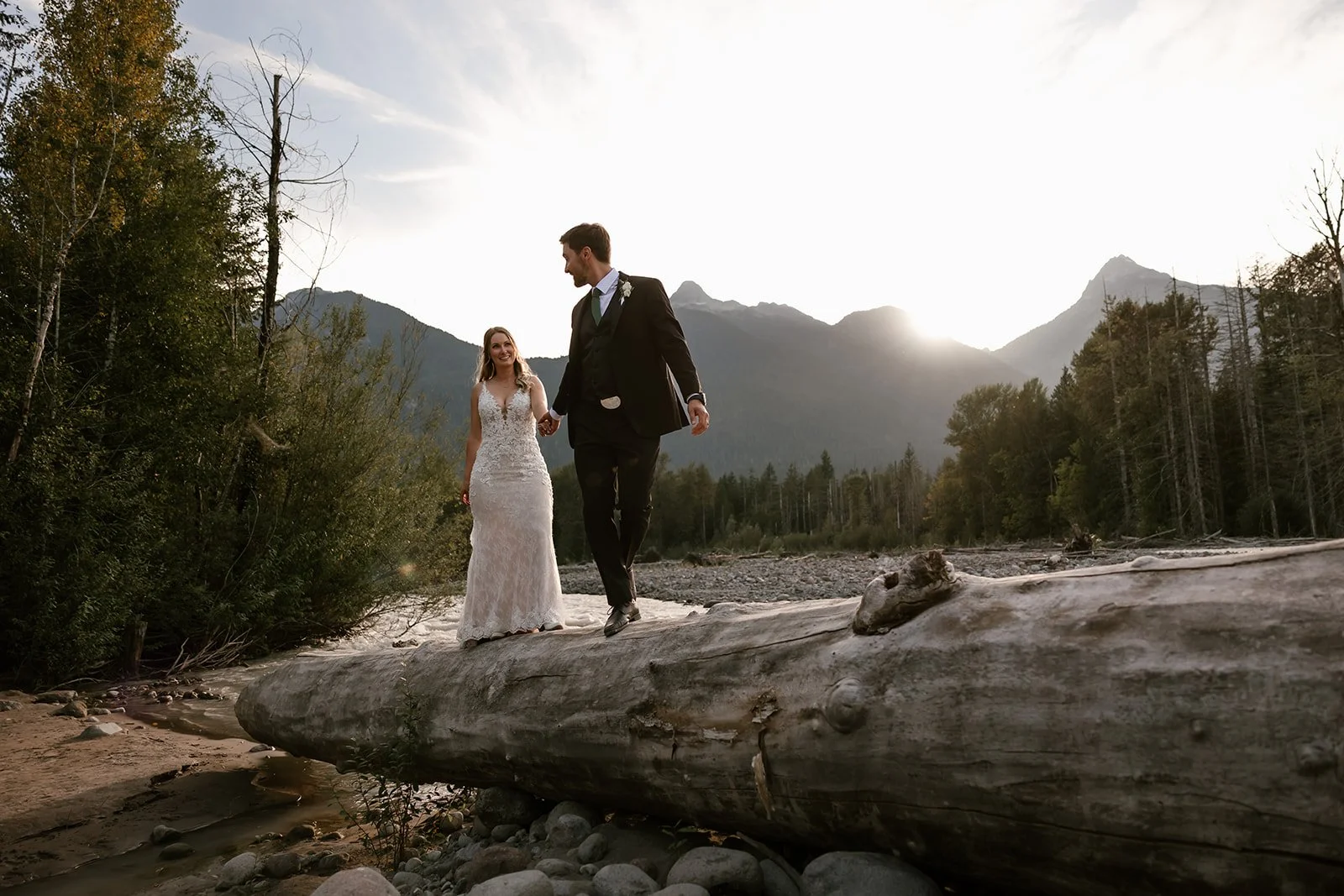  A bride and groom standing on a log with the the forest, mountains and river behind them.  