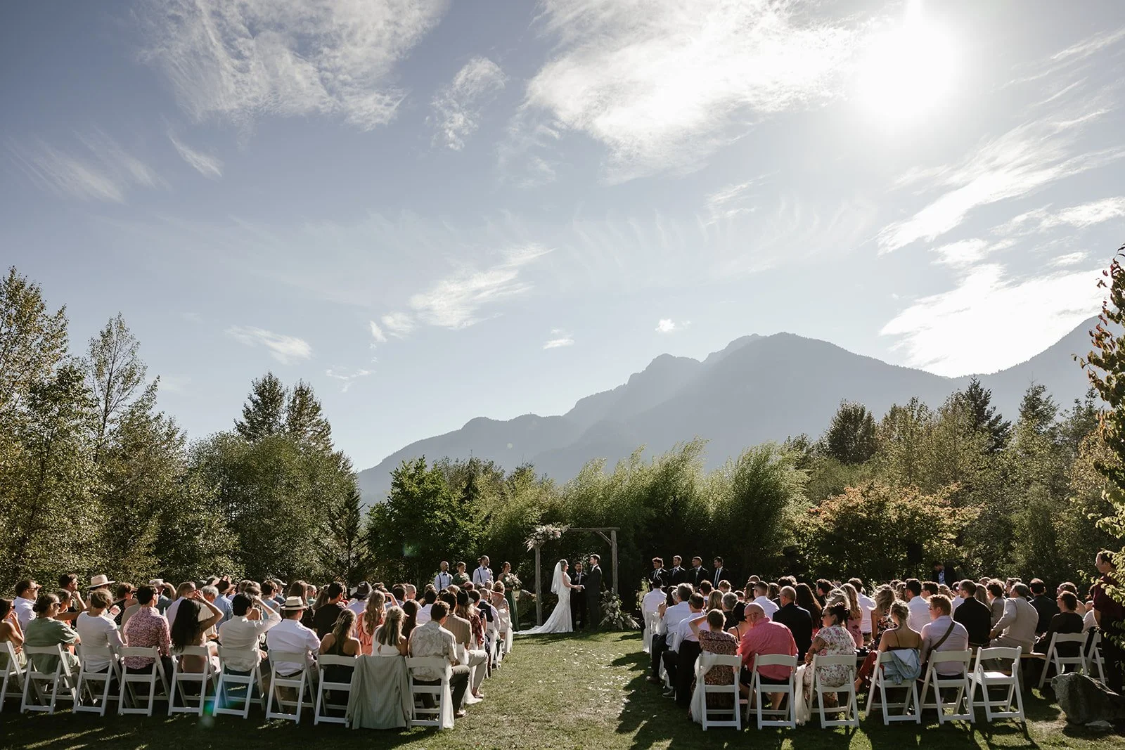  A wedding ceremony in a beautiful outdoor setting with guests seated on either side, the couple in the middle of the aisle, and trees and mountains in the background.  