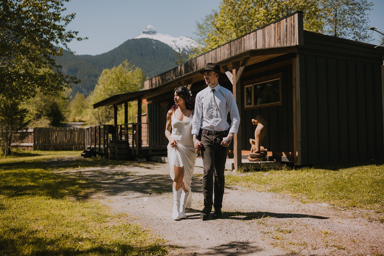  A bride and groom in cowboy boots and a cowboy hat in front of a rustic cabin with mountains behind them.  