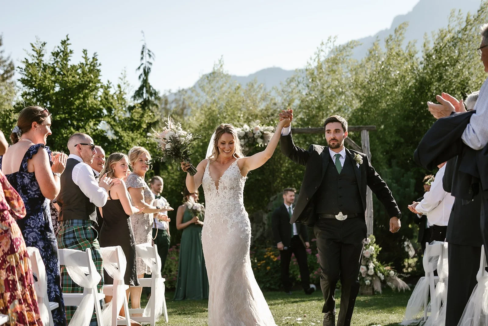  A bride and groom walking down the aisle while holding hands with big smiles and guests on either side, with mountains and greenery behind them.  