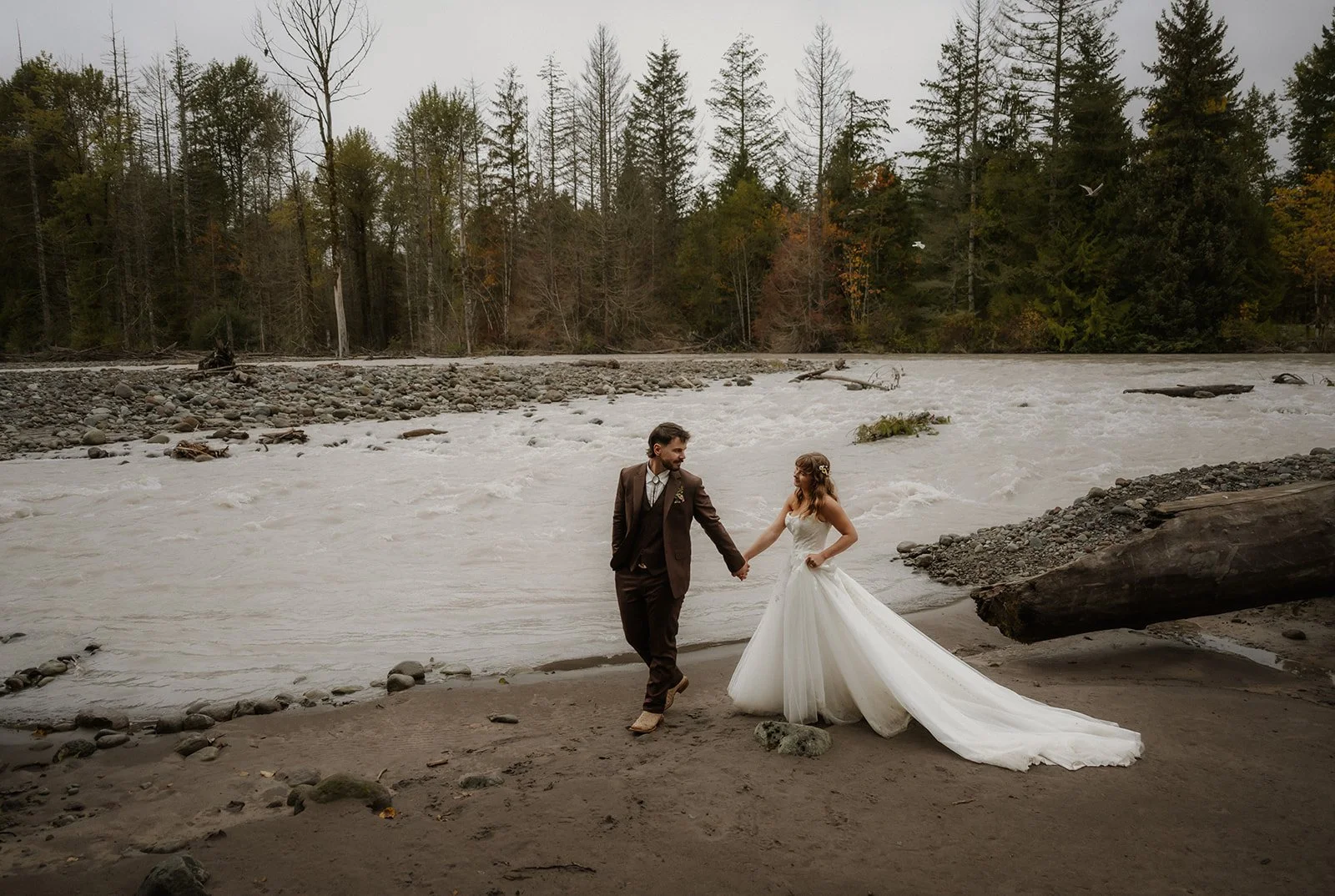  A bride and groom walking down a sandy beach, holding hands, with a river and trees behind them.  