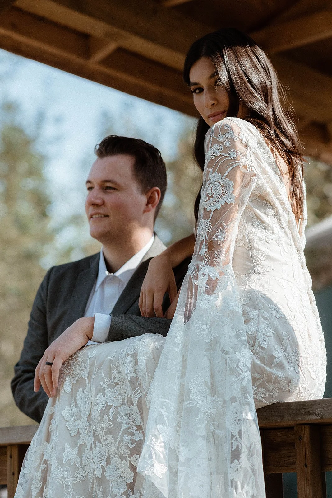  A bride sitting in a lace white dress with long sleeves with her groom behind her looking to the side.  