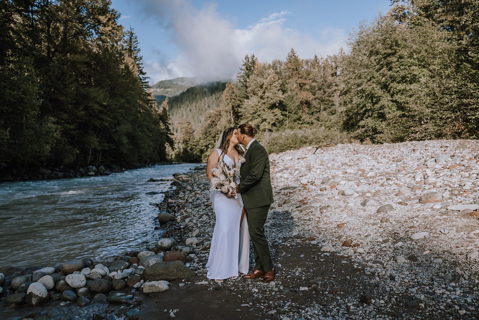  A bride and groom holding each other on a rocky beach with a river and trees and blue sky behind them.  