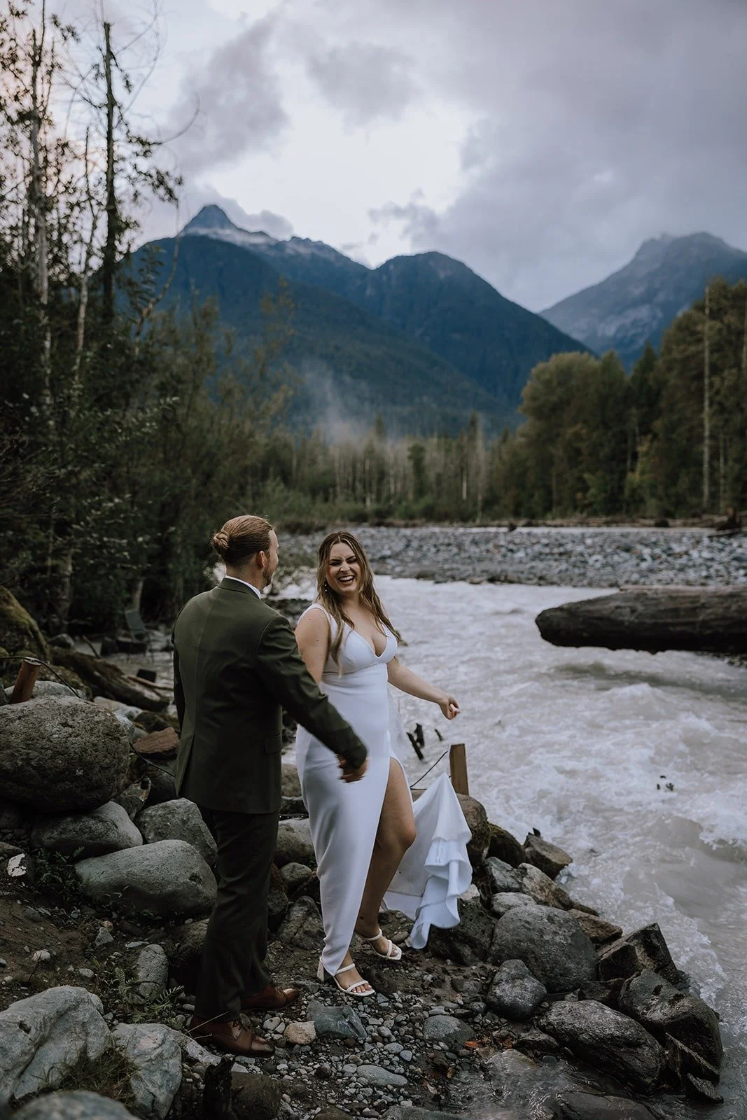  A bride and groom on a river side with mountains behind them.  