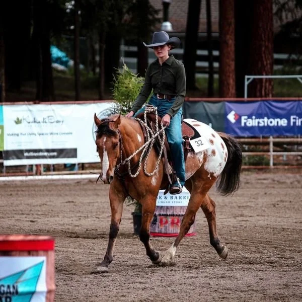 A woman riding an Appaloosa brown and white horse in a cowboy hat.