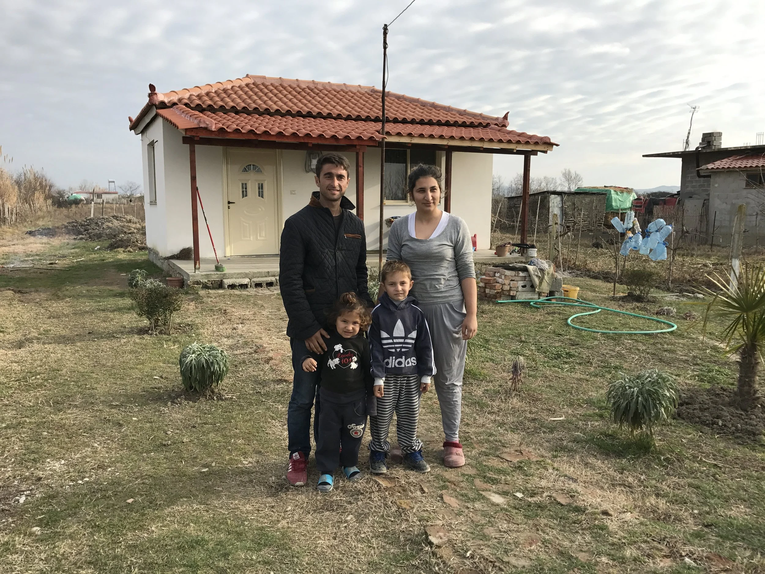 A family of five standing outdoors in front of a small house with a red-tiled roof. The family includes two adults and three children. The background shows a rural setting with trees, other houses, and a cloudy sky.