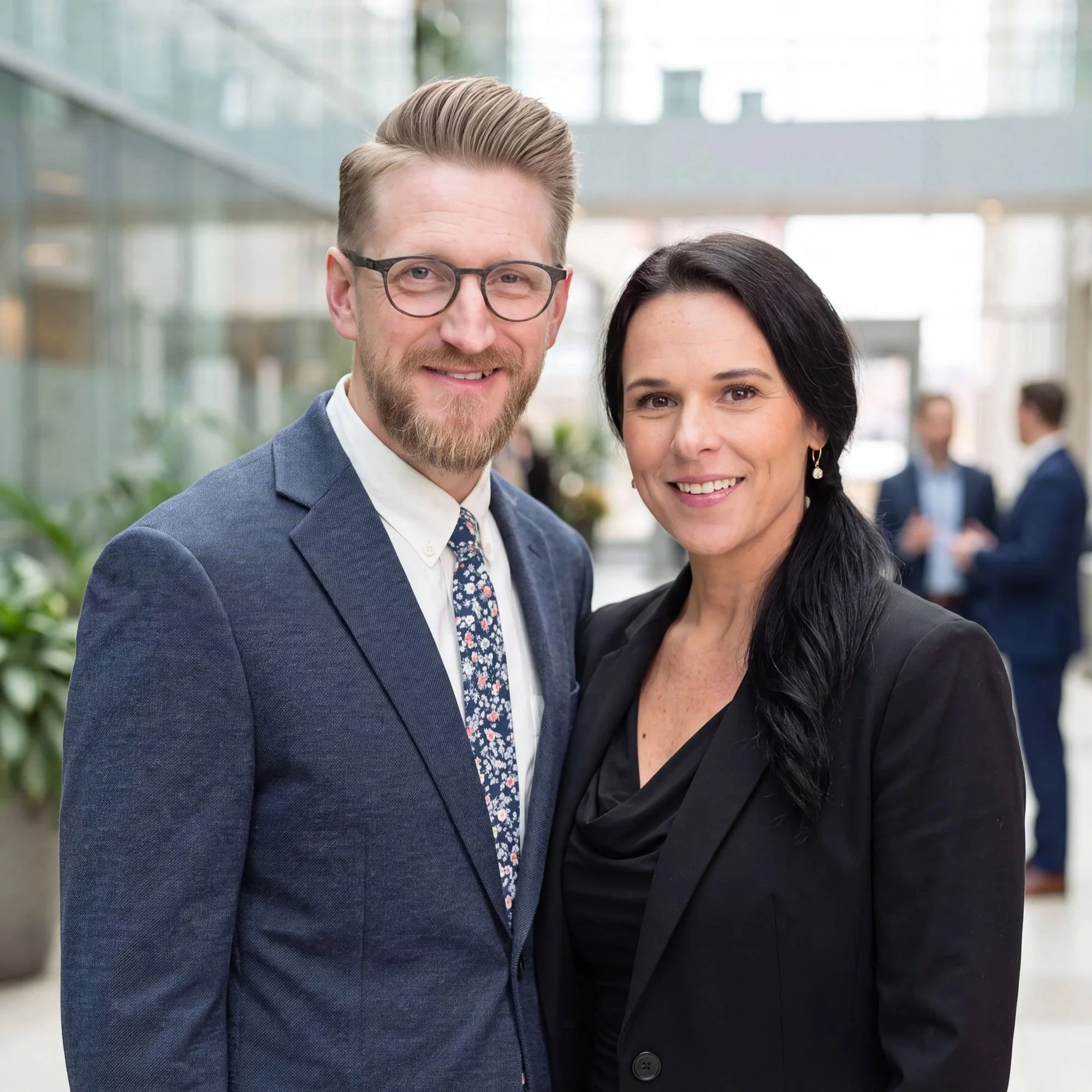 Smiling man with glasses and beard, wearing a blue suit and tie, standing next to a woman with dark hair, both dressed in professional attire, in a bright modern office environment.