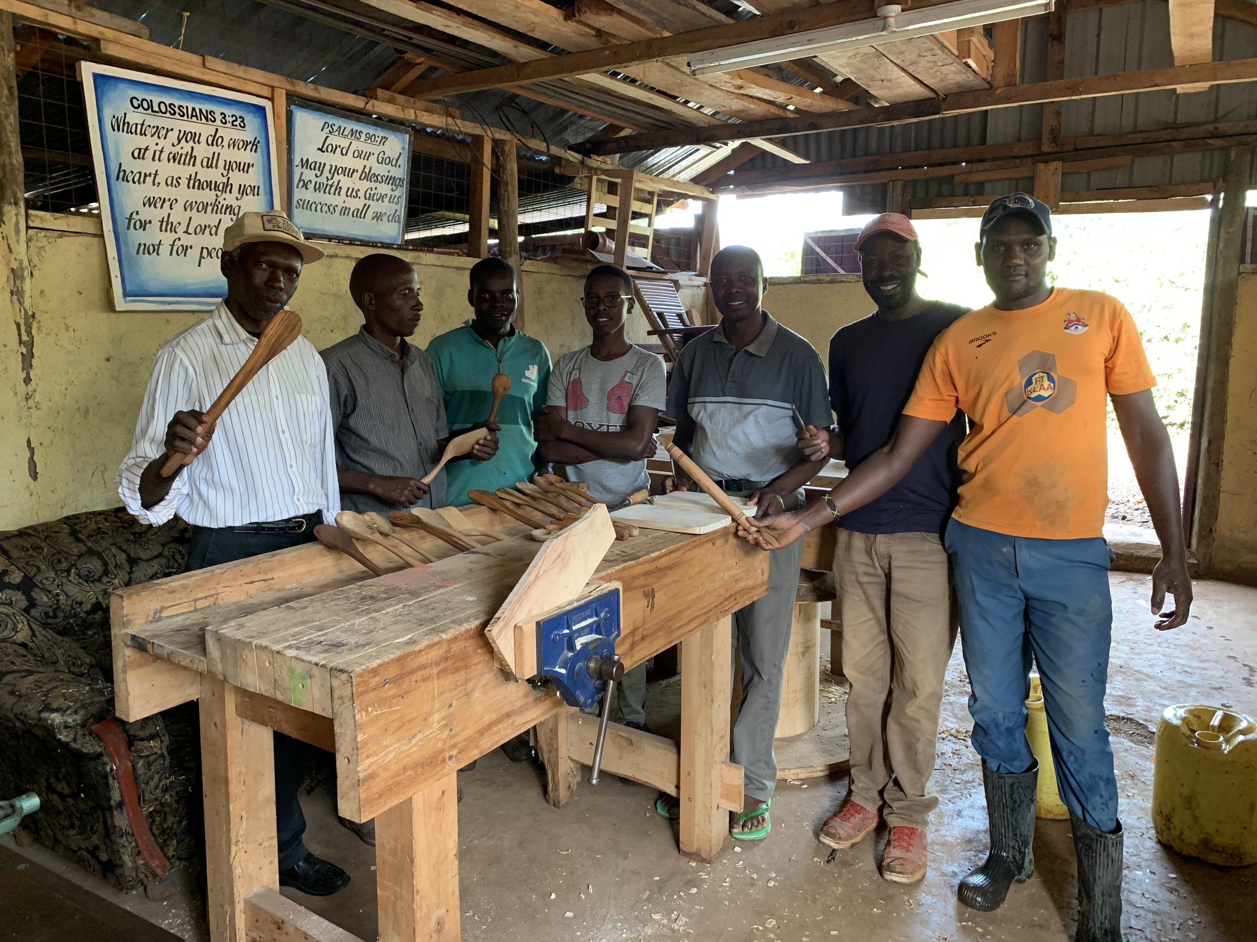 Group of seven people standing behind a wooden workbench with woodworking tools inside a workshop, with signs containing Bible verses on the wall behind them.