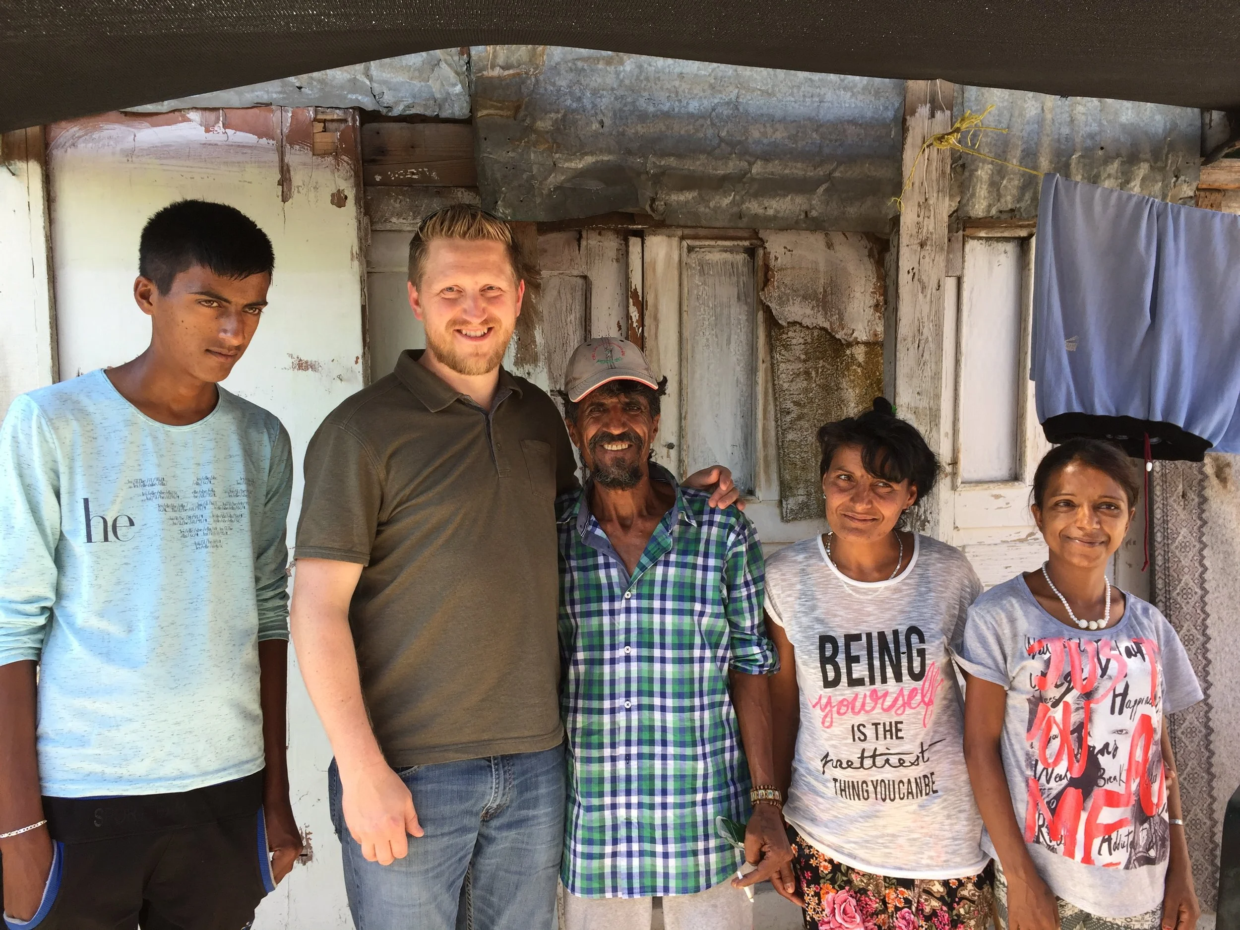 A group of six people standing outdoors in front of a weathered house with wooden and metal exterior walls, smiling with some holding hands.
