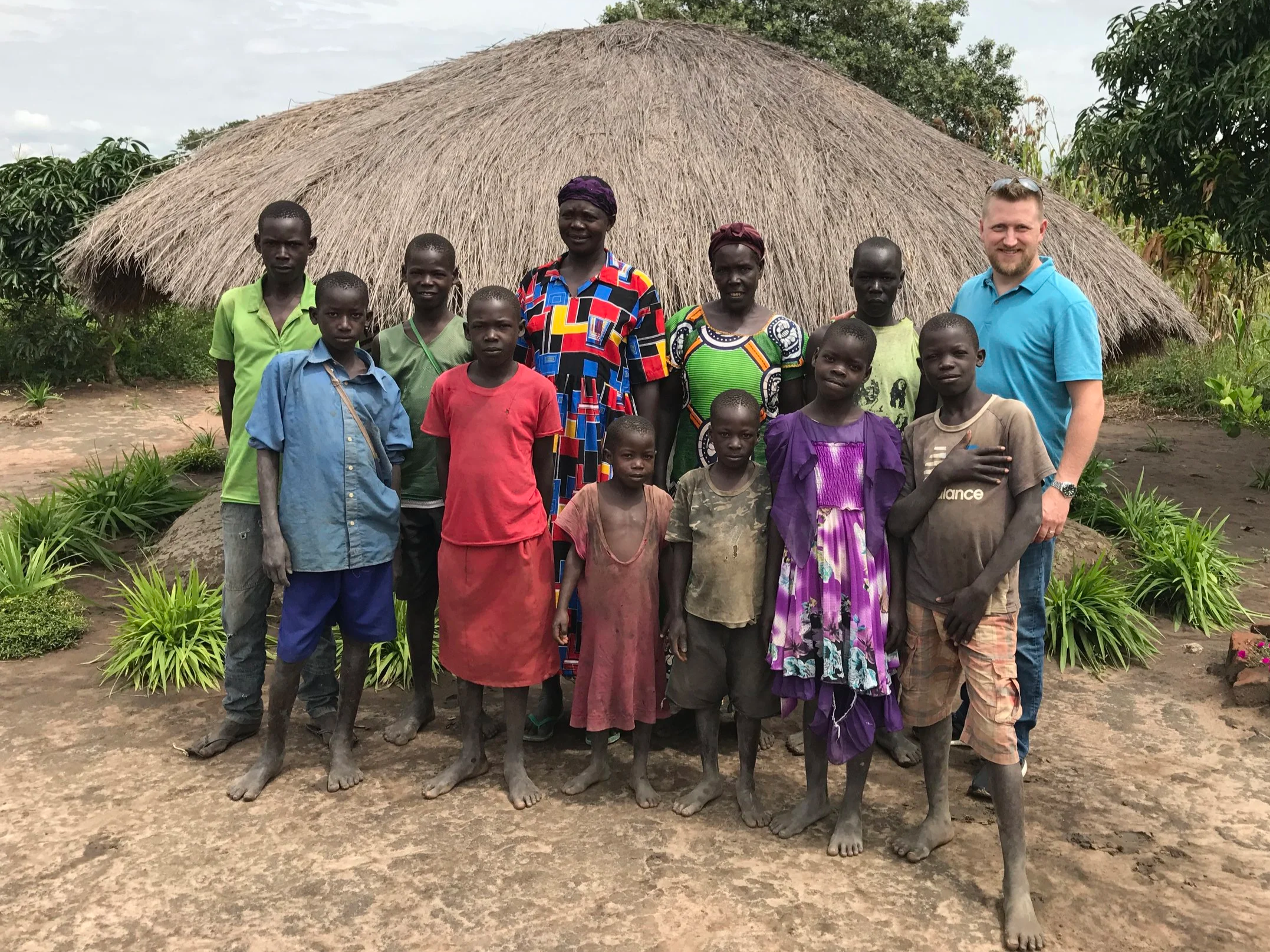 A group of fifteen people, including children and adults, standing in front of a thatched-roof hut surrounded by lush greenery, in a rural setting.