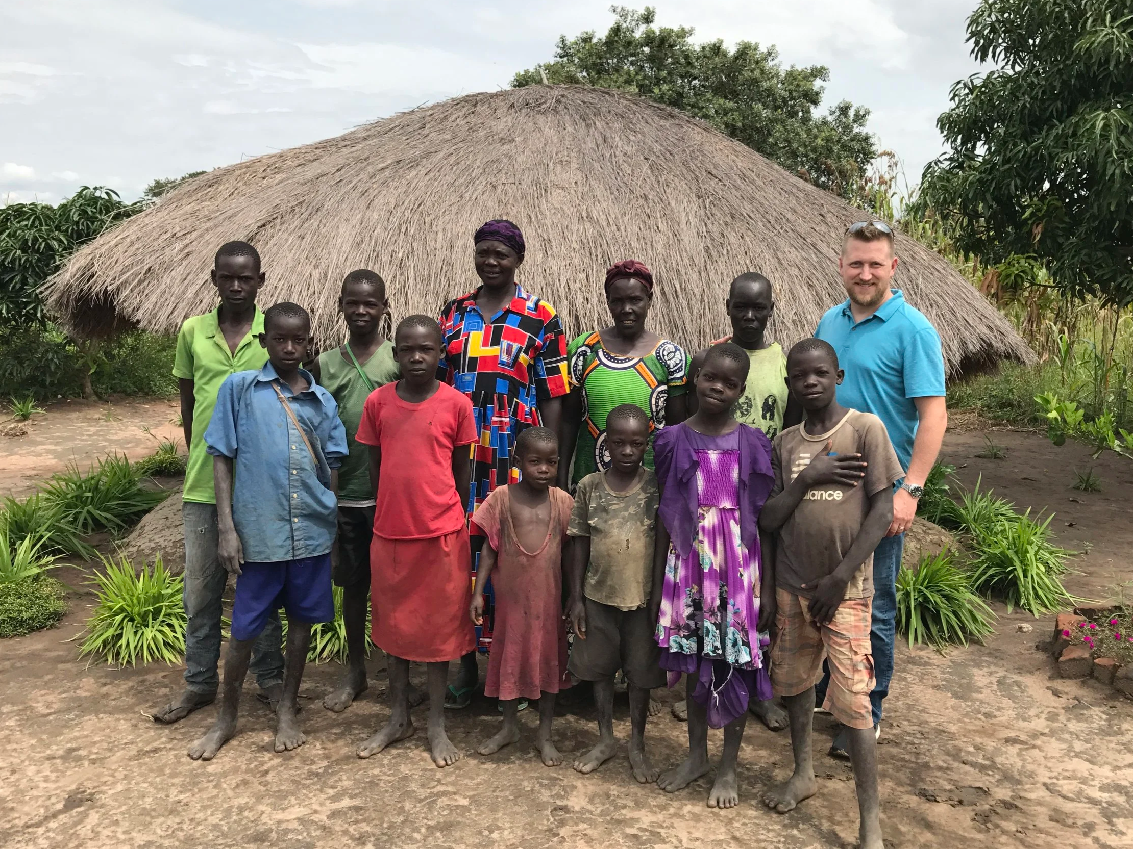 A group of people, including children and adults, standing in front of a traditional thatched-roof hut, with greenery and flowering plants around them, on a dirt ground under a cloudy sky.