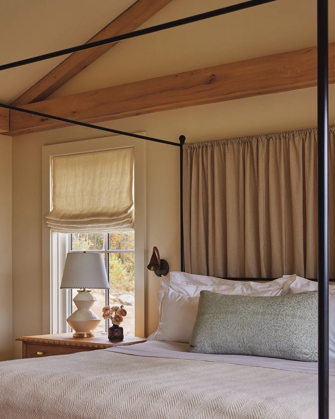 Wooden beams and layers of texture add depth to the primary bedroom at our #LakeWaramaug project. The back of the four-posted bed is hung with &lsquo;Little Stripe&rsquo; by @nicholasherbertltd, which, together with a layering of brass wall lights, a