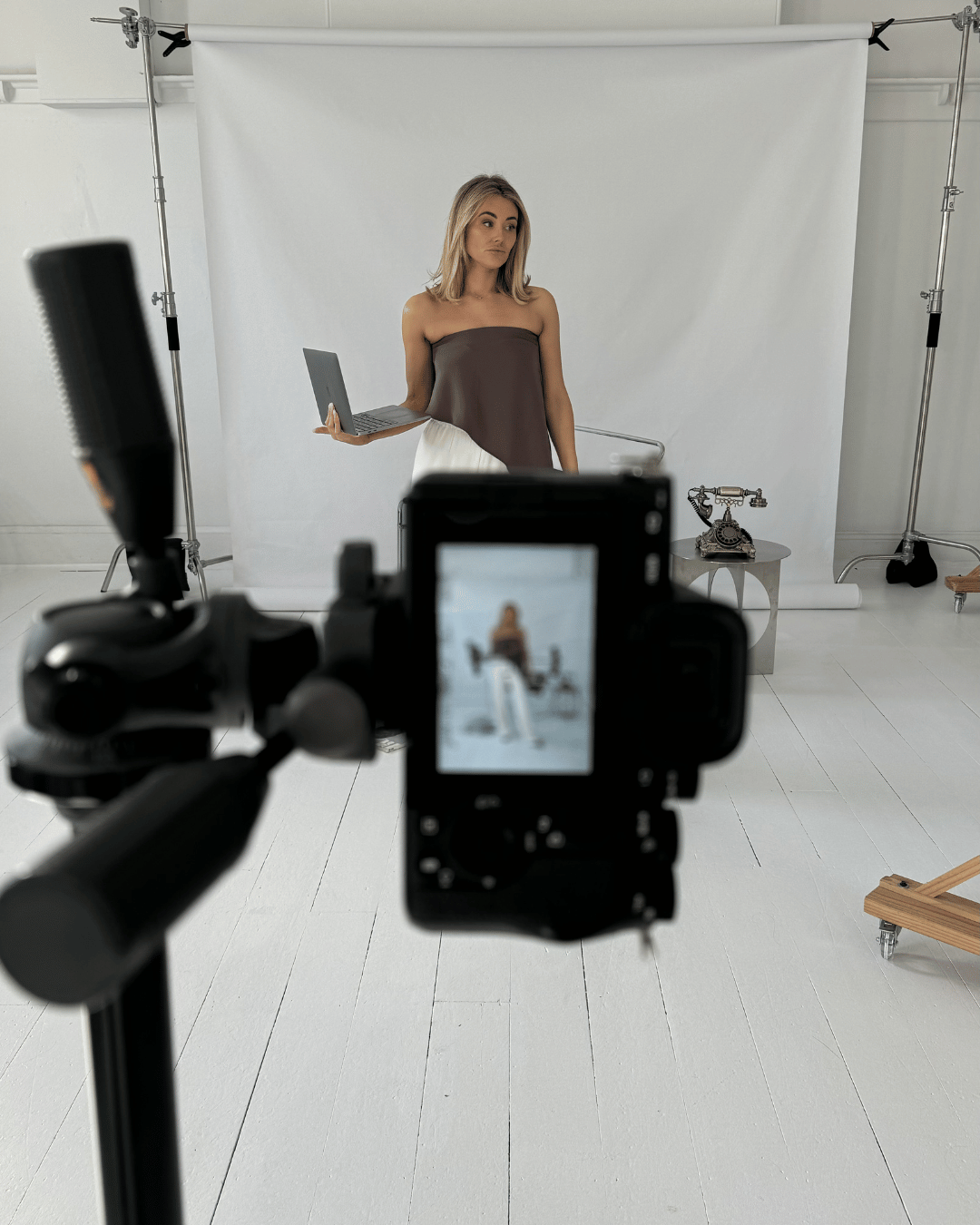 A woman stands in front of a white backdrop, holding a laptop in one hand. A camera is focused on her, capturing her pose, with studio equipment and a vintage telephone on a table nearby.