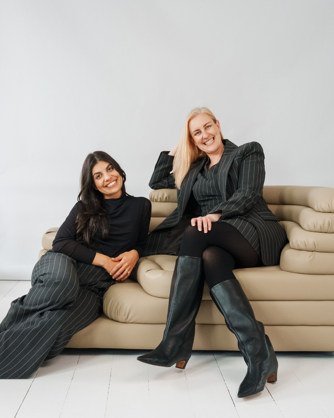 Two women smiling and posing on a beige sofa against a plain white wall; one sitting on the floor and the other sitting on the sofa.