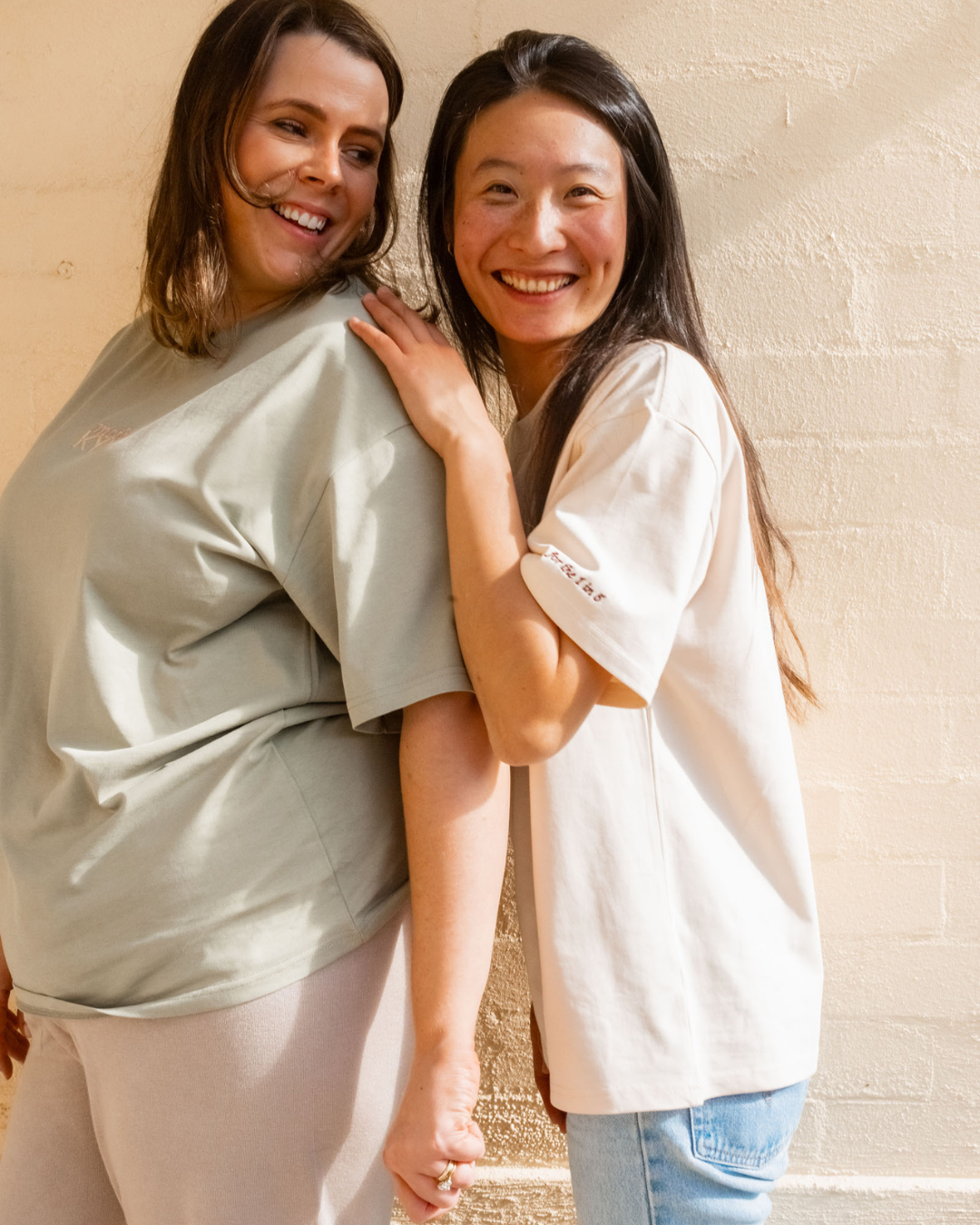 Two women smiling and hugging, standing against a beige wall.