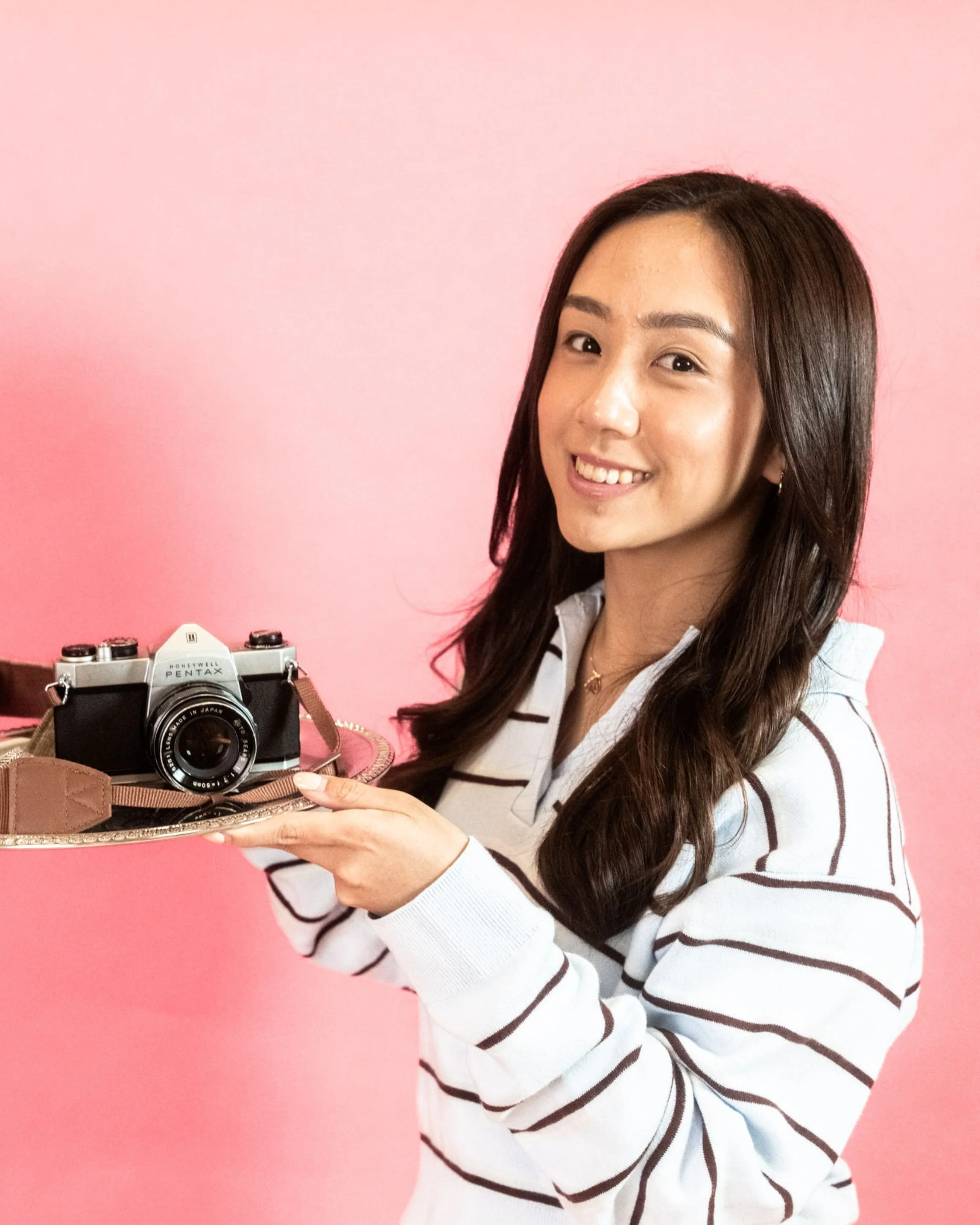 A young woman with long dark hair smiling while holding a vintage camera on a tray against a pink background.