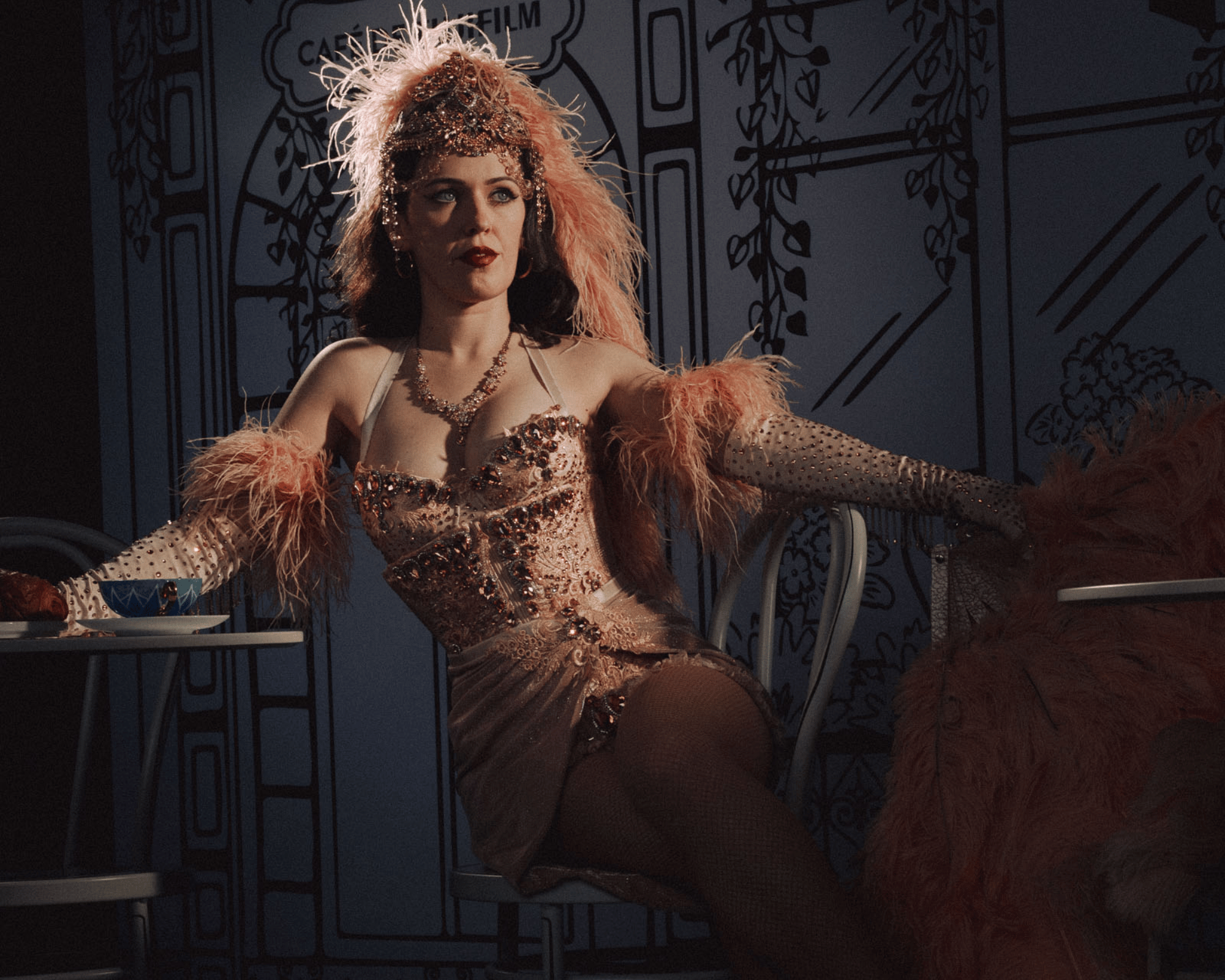 A woman dressed in glamorous, vintage-style attire with intricate beadwork, a feathered headpiece, and dark lipstick sitting at a table in a dimly lit setting.