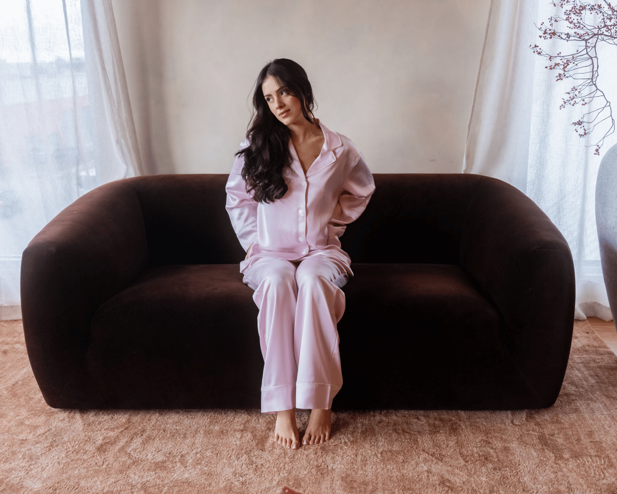 A woman sitting on a brown sofa in a pink silk pajama set in a room with sheer white curtains, a decorated wall, and a beige carpet.
