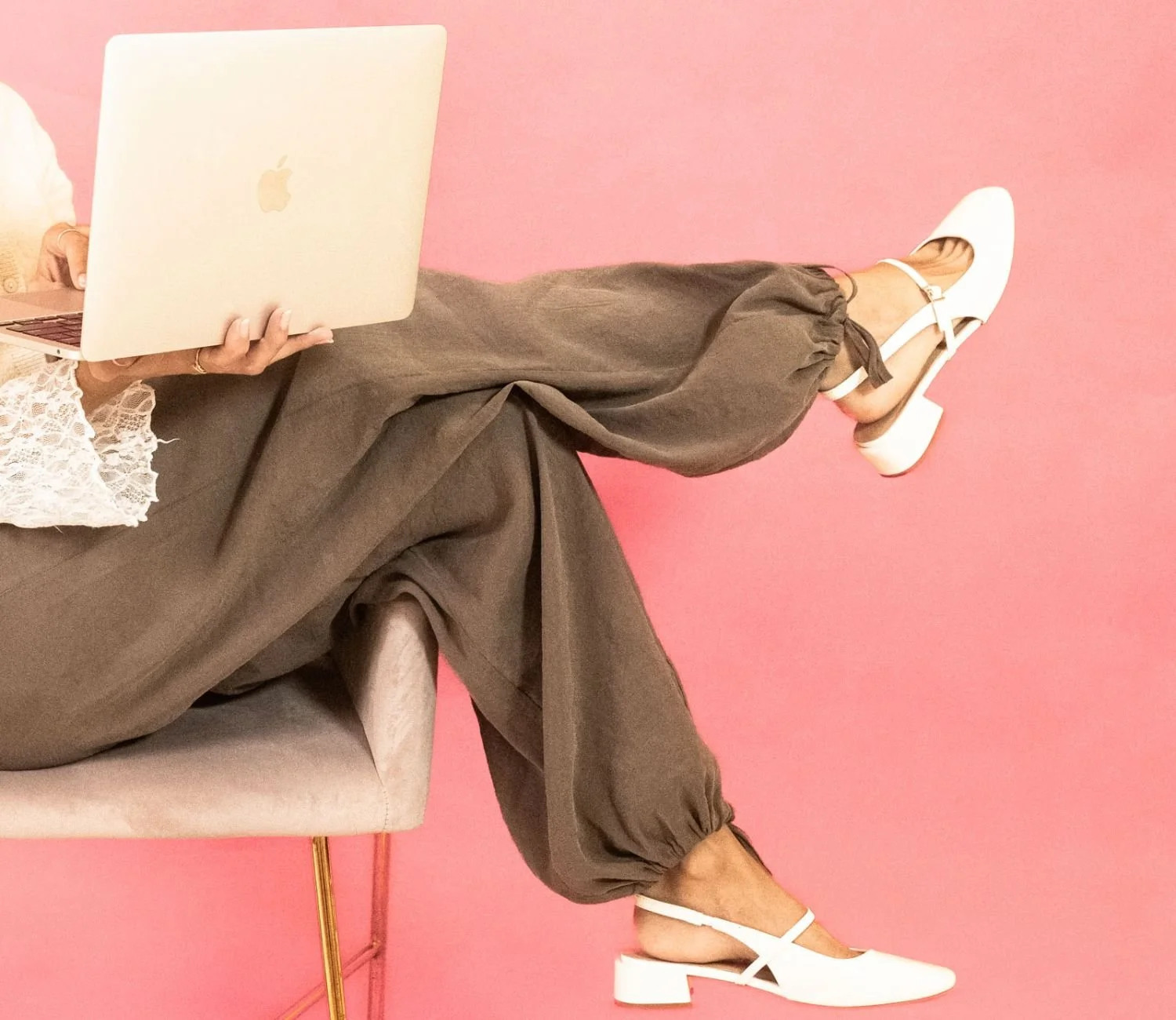 Person sitting on a beige chair against a pink background, wearing brown pants, white high-heeled shoes, and working on a silver laptop.