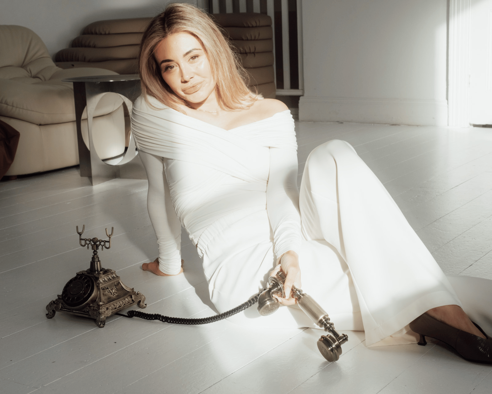 A woman with shoulder-length blonde hair, dressed in a white off-shoulder dress, is sitting on a white wooden floor in a bright room with white walls. She is holding a vintage black rotary telephone receiver and a silver candle holder. The vintage te