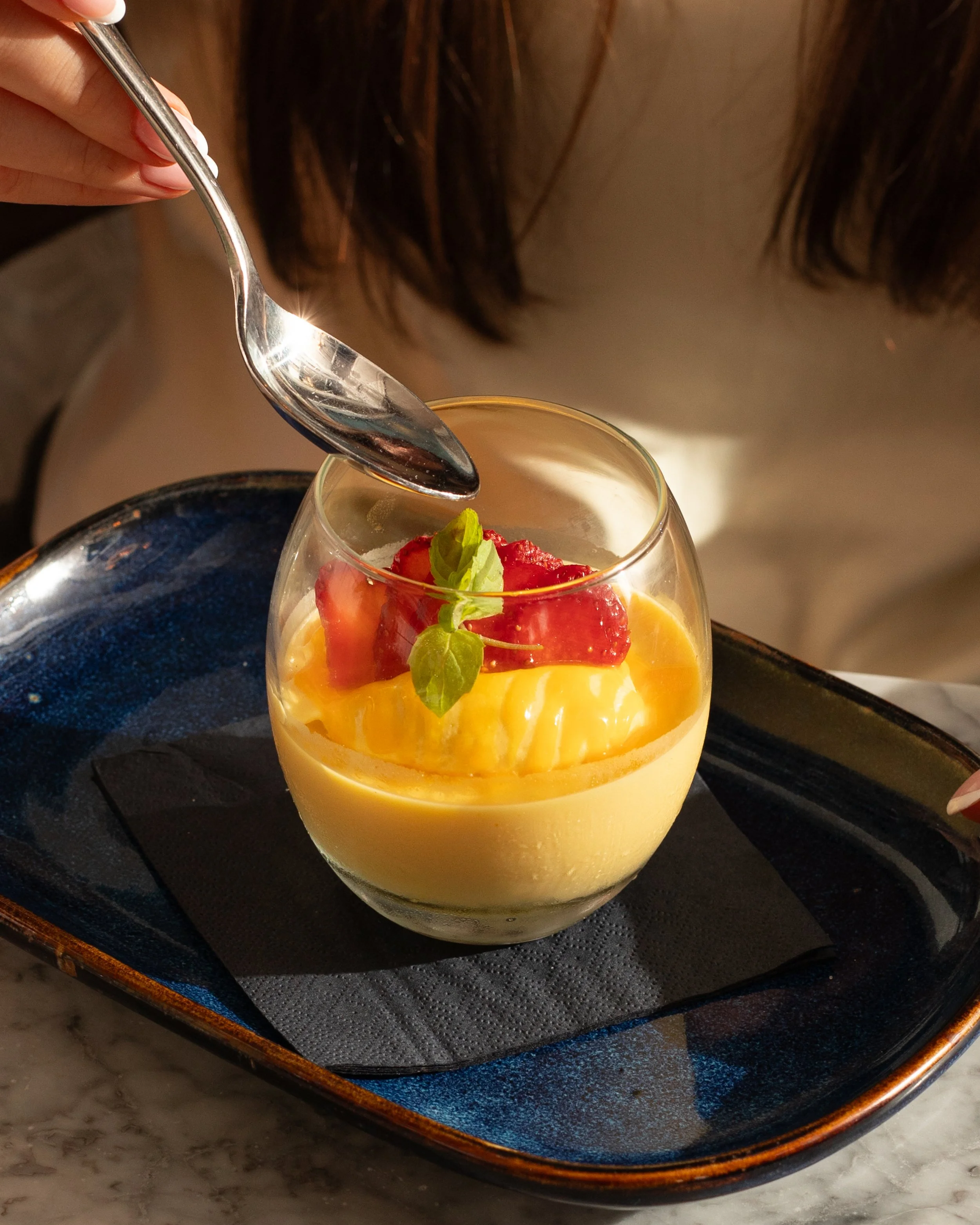 A glass of yellow dessert topped with strawberries and green garnish, placed on a black plate with a dark blue rim