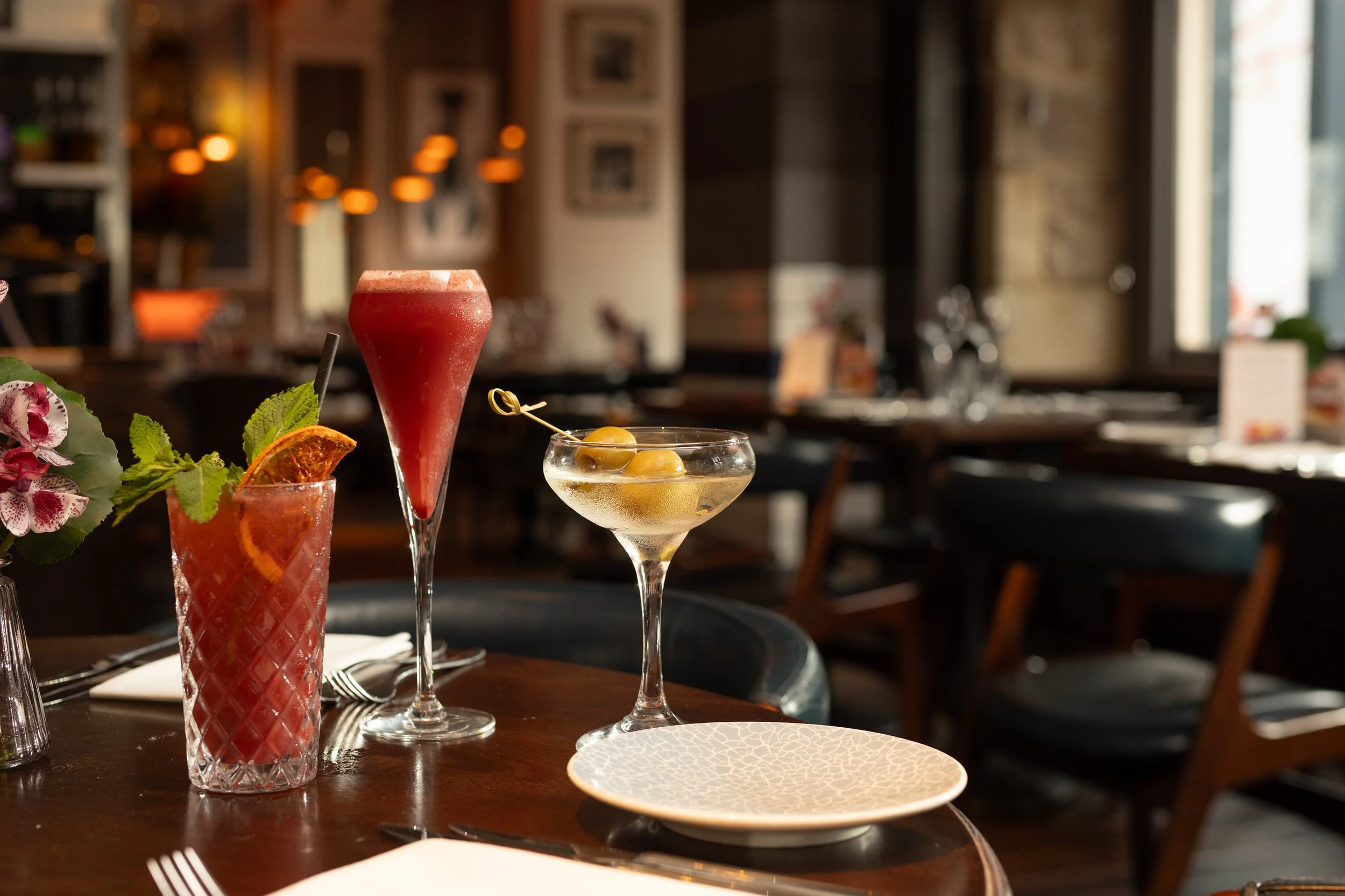 A variety of cocktails on a restaurant table, including a pink drink with mint and orange, a red drink in a tall glass, a martini with olives, and a straw in another glass, with a blurred restaurant interior background.