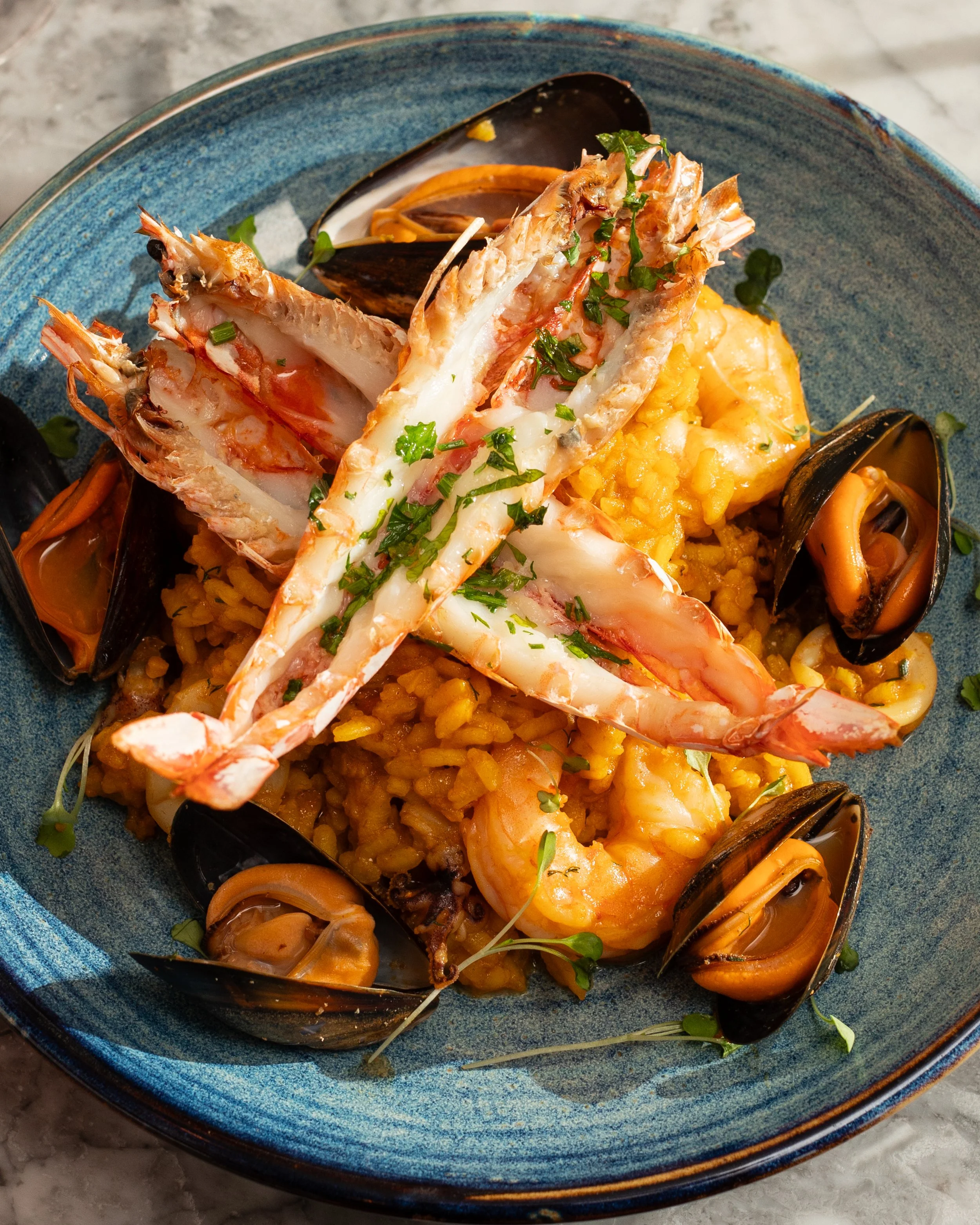 Seafood paella with large shrimp, mussels, and rice, garnished with chopped herbs served on a blue ceramic plate.
